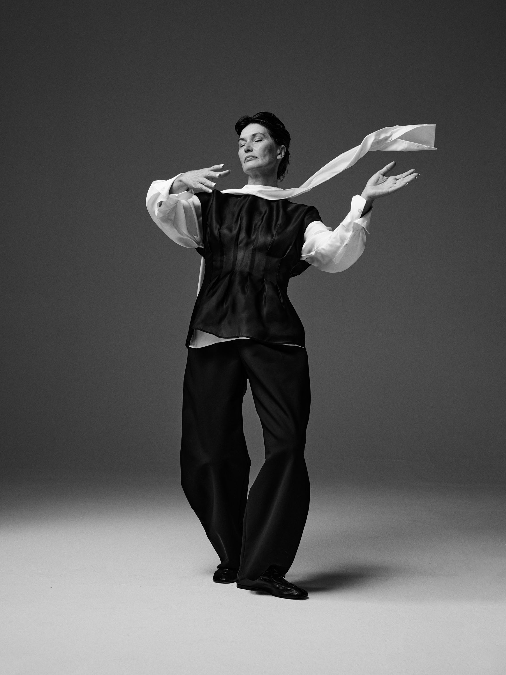Woman wearing a cream scarf blouse, organza waisted top and black trousers standing against a plain background