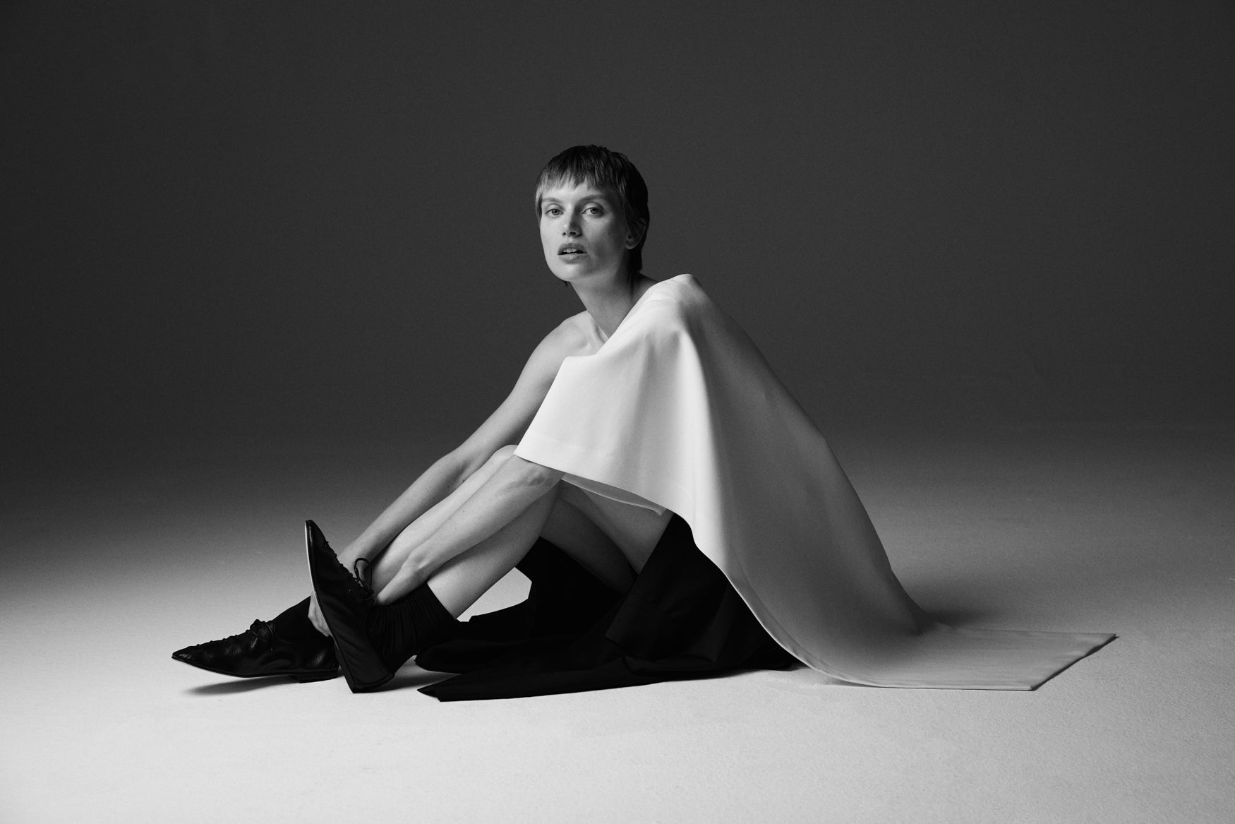 Woman sitting down on the ground wearing a cream cape top draping over the ground against a plain background