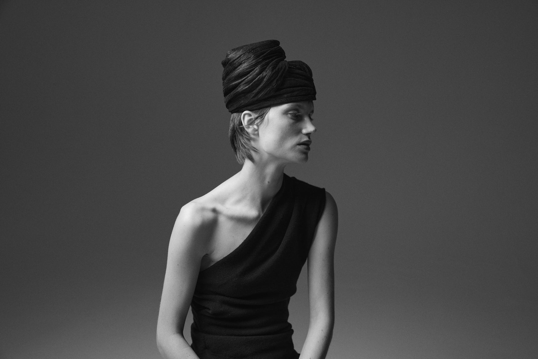 Black and white portrait of a woman wearing a black one shoulder top and an organza pillbox hat