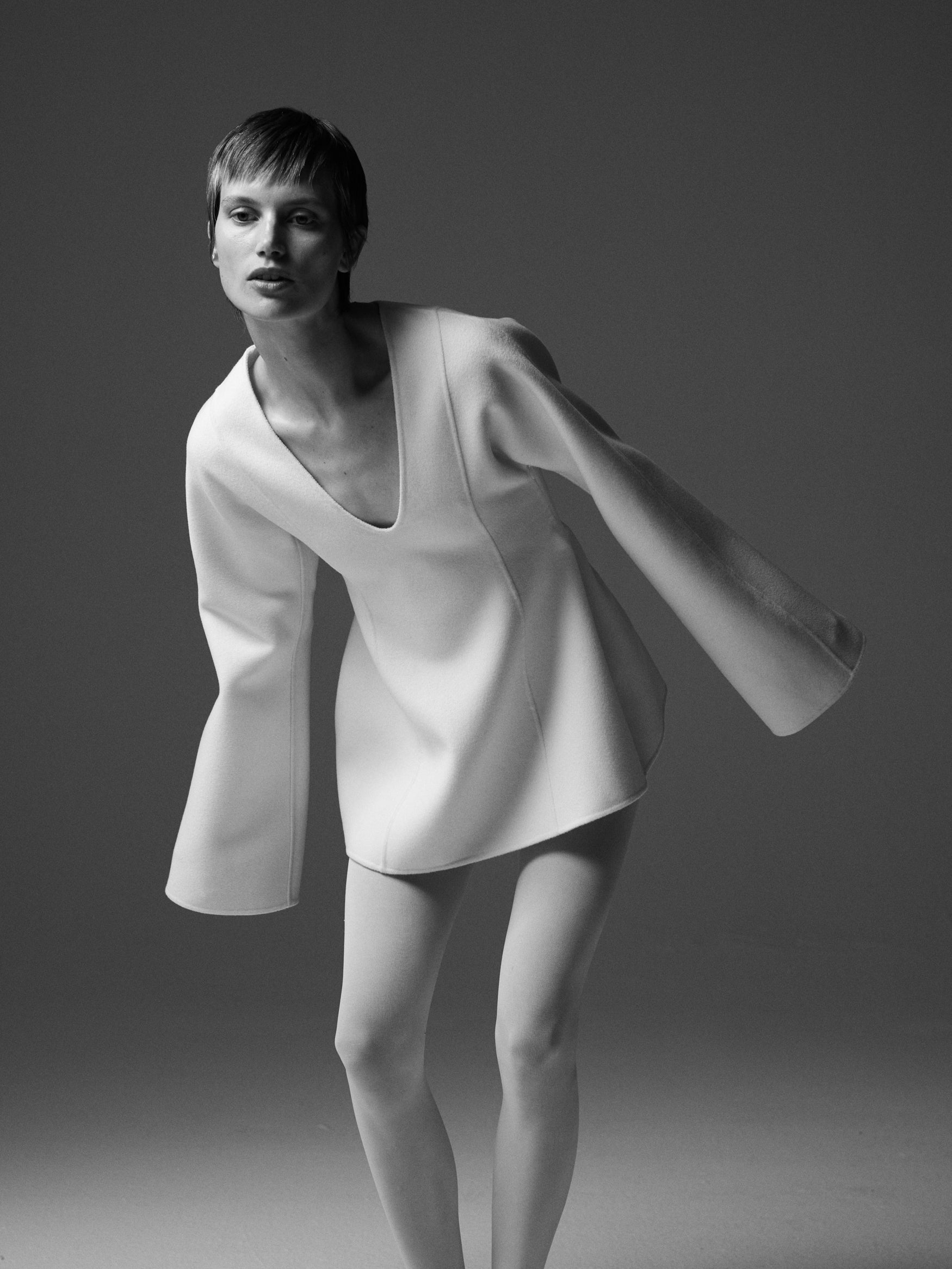 Black and white image of a woman leaning forward in a cream double faced sculptural top with v-neck against a plain backdrop