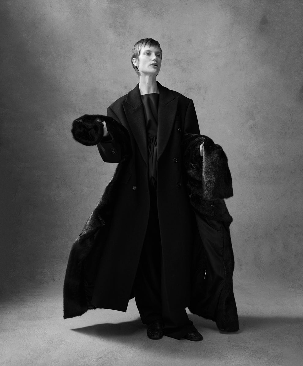 Woman wearing a long black coat, holding a faux fur coat around her waist 