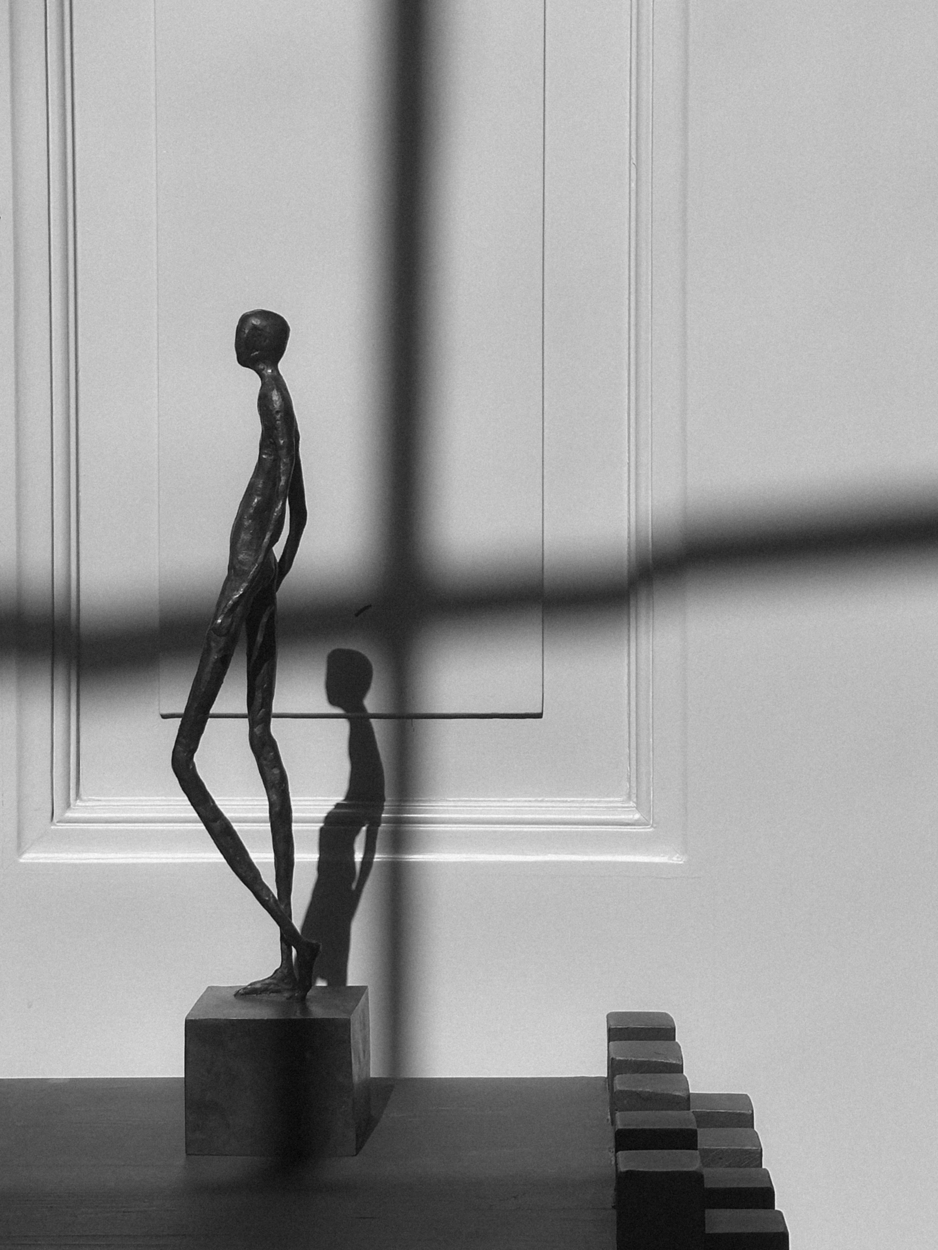 Abstract sculpture of an elegant figure on a pedestal against a white wall with shadows casting the wall