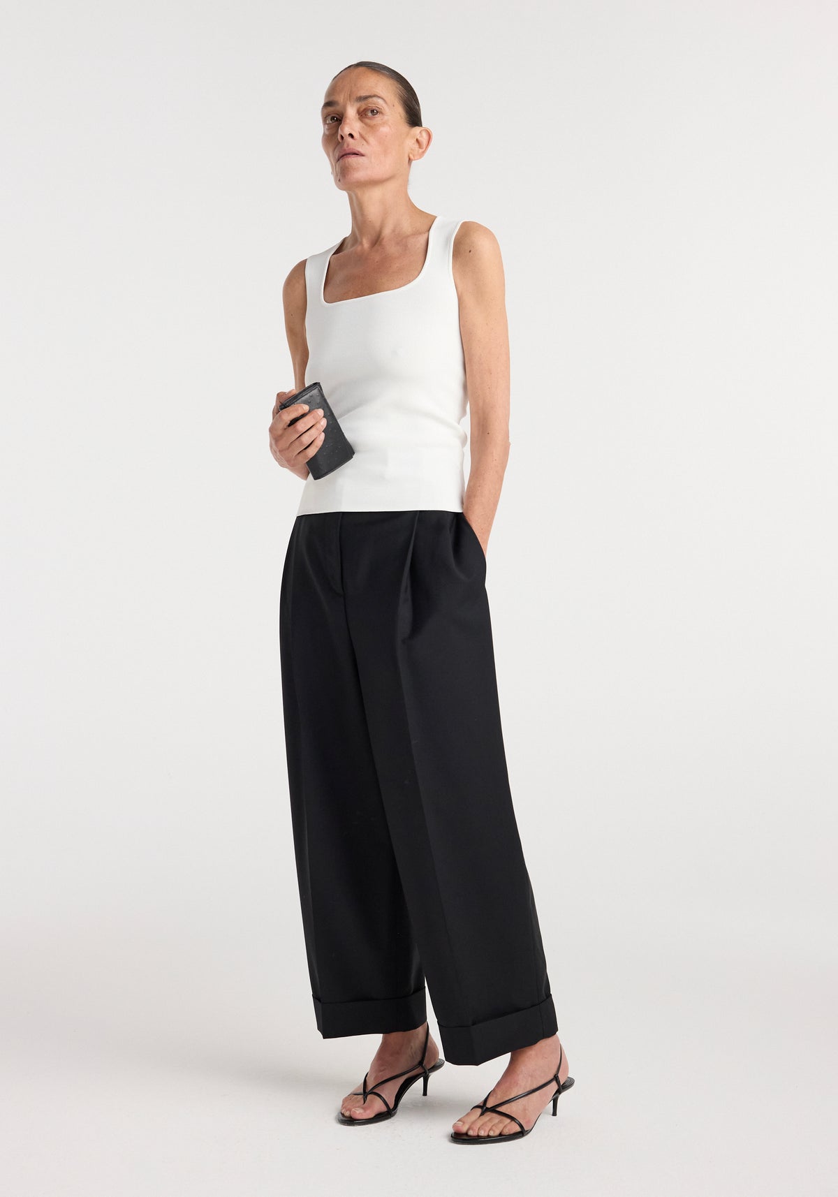 Woman wearing a bustier shaped knitted top | optic white paired with wide-leg black trousers and black strappy heels, holding a small black clutch against a plain background.