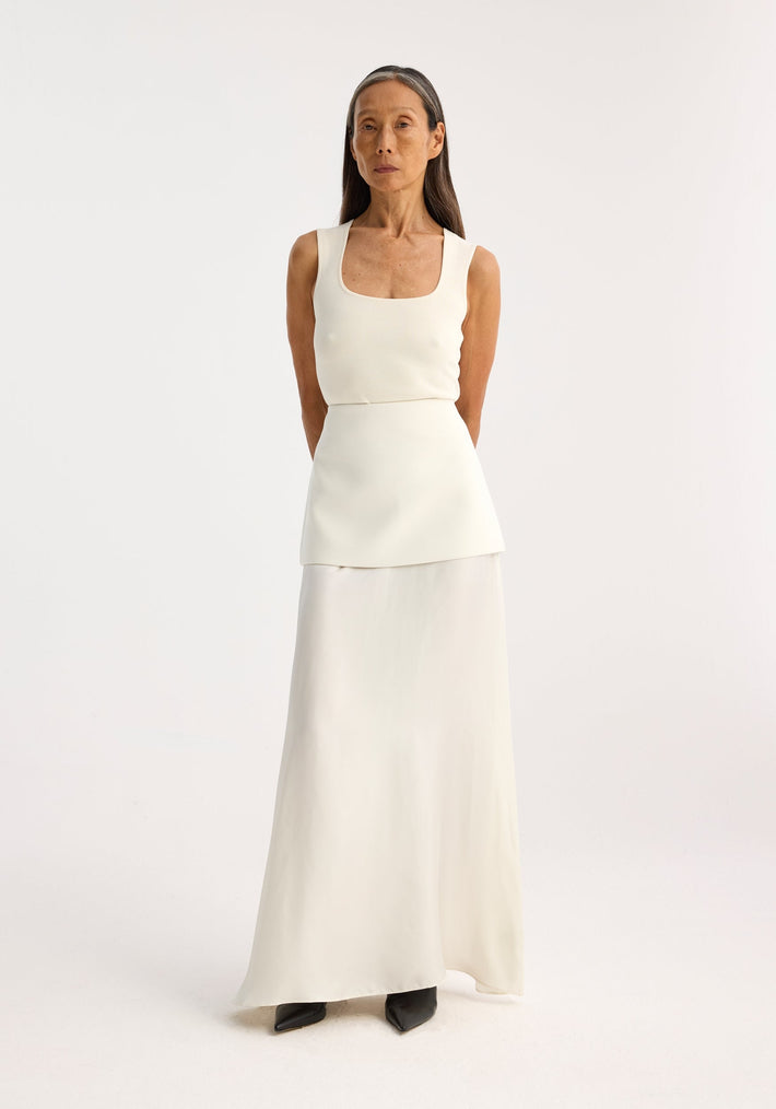 Person in a bustier-shaped knitted top, off white, with a square neckline, paired with a layered skirt. Arms behind back, neutral expression.