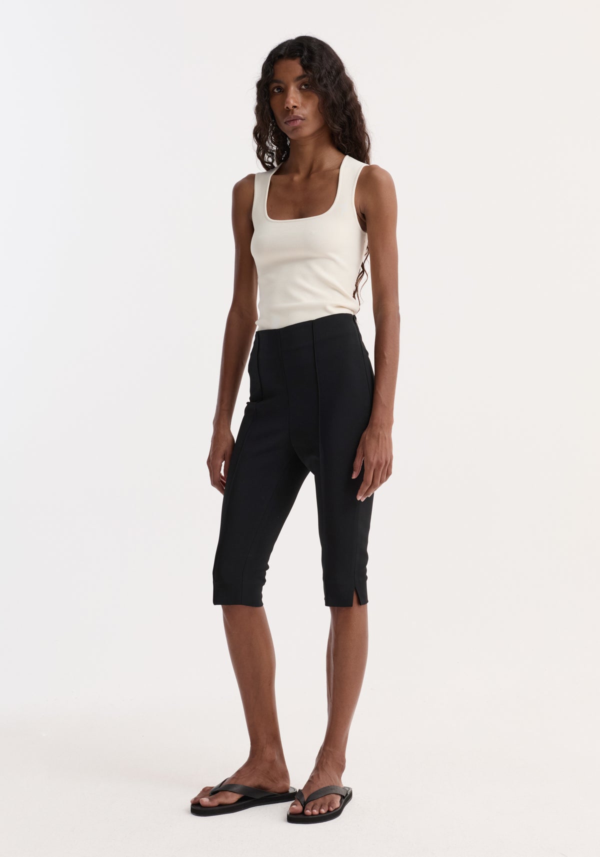 Woman wearing a sleeveless Bustier shaped knitted top | off white paired with black cropped pants and black flip-flops standing against a plain white background.