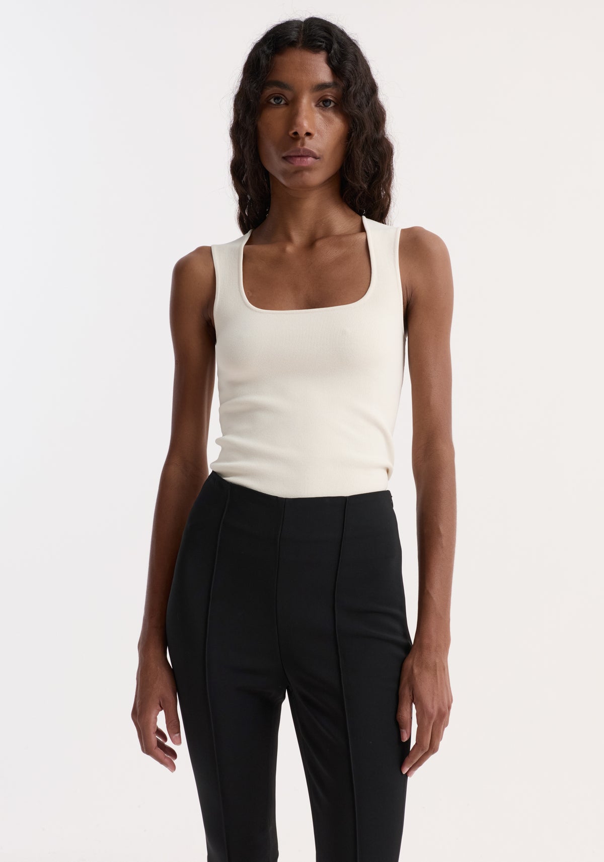 Woman wearing an off-white Bustier shaped knitted top | off white with a square neckline and sleeveless design, paired with high-waisted black pants against a white background.