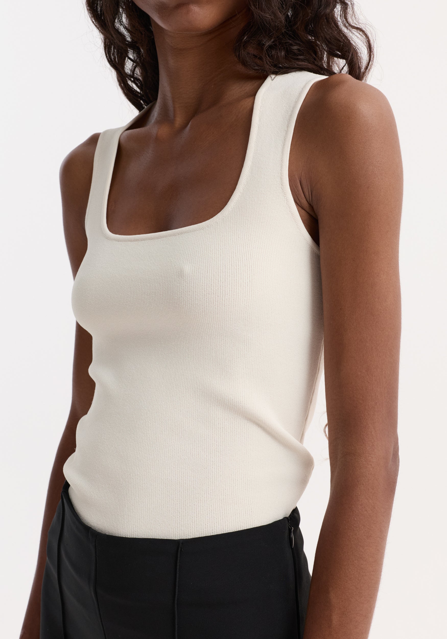 Woman wearing a fitted Bustier shaped knitted top | off white with a square neckline paired with black pants against a white background.