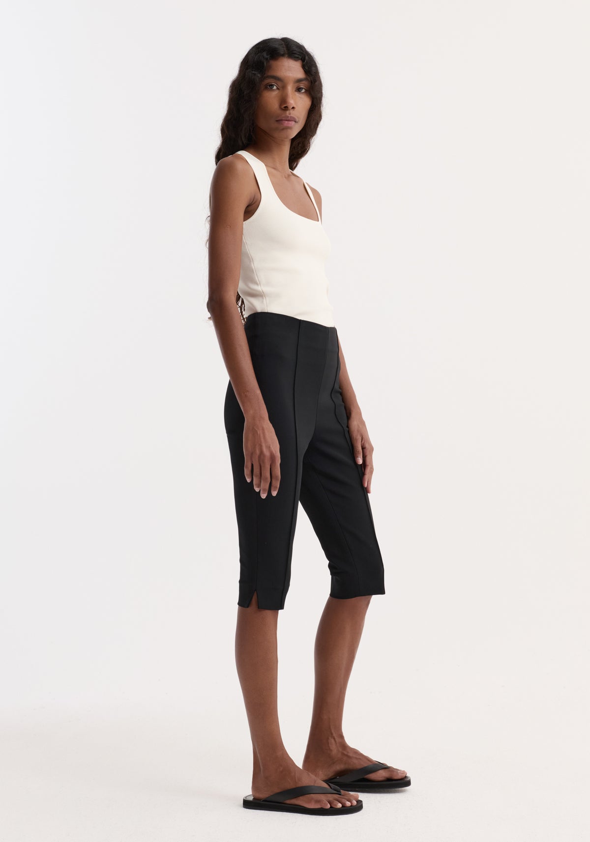 Woman wearing a sleeveless bustier shaped knitted top | off white paired with fitted black cropped pants and black sandals, standing in profile against a white background.