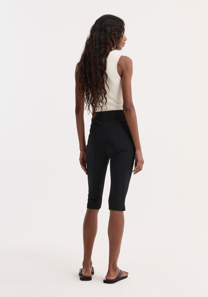 Woman wearing a Bustier shaped knitted top in off white, paired with knee-length black pants and black slide sandals, showcasing a fitted, sleeveless style from behind.