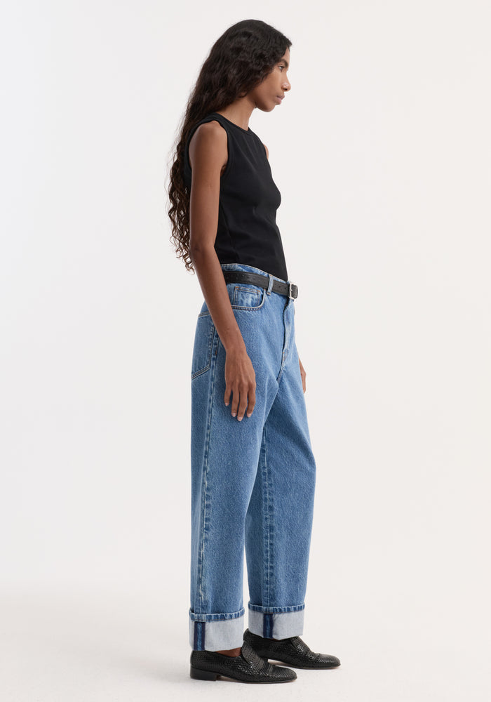 Side-profile of a woman wearing a Dense rib tank top in black, styled with high-waisted jeans and loafers, highlighting its fitted, ribbed design.