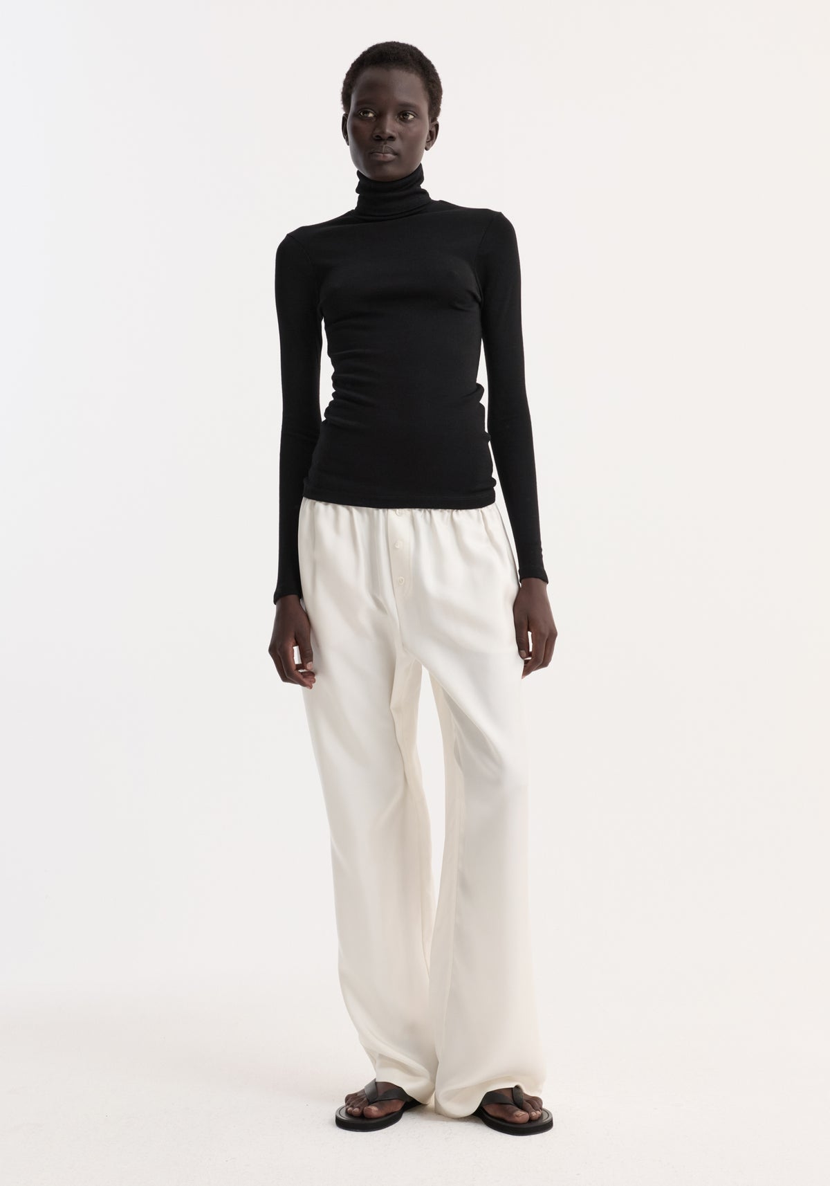 Model wearing a fine merino turtleneck | black paired with loose white pants and black sandals, standing against a plain light background.