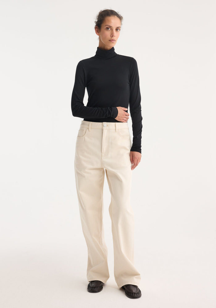 Person wearing a Fine merino turtleneck in black, fitted, long-sleeve design, paired with high-waisted trousers and loafers, showcasing a minimalist, chic style.