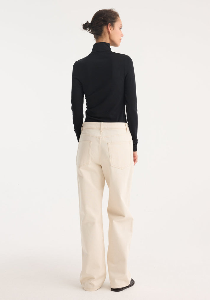 Person wearing a fitted Fine merino turtleneck in black, styled with high-waisted cream pants and black loafers, hair in a bun, against a plain background.