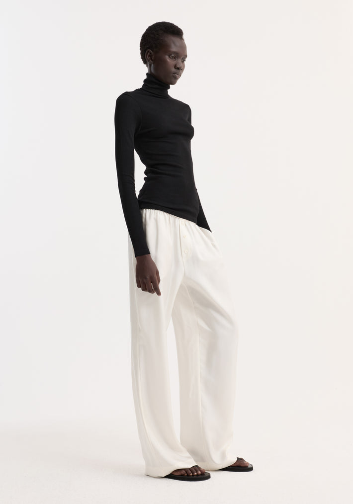 Model in Fine merino turtleneck | black, showcasing a fitted turtleneck with long sleeves, paired with wide-leg trousers; highlights the turtleneck's sleek, versatile design.