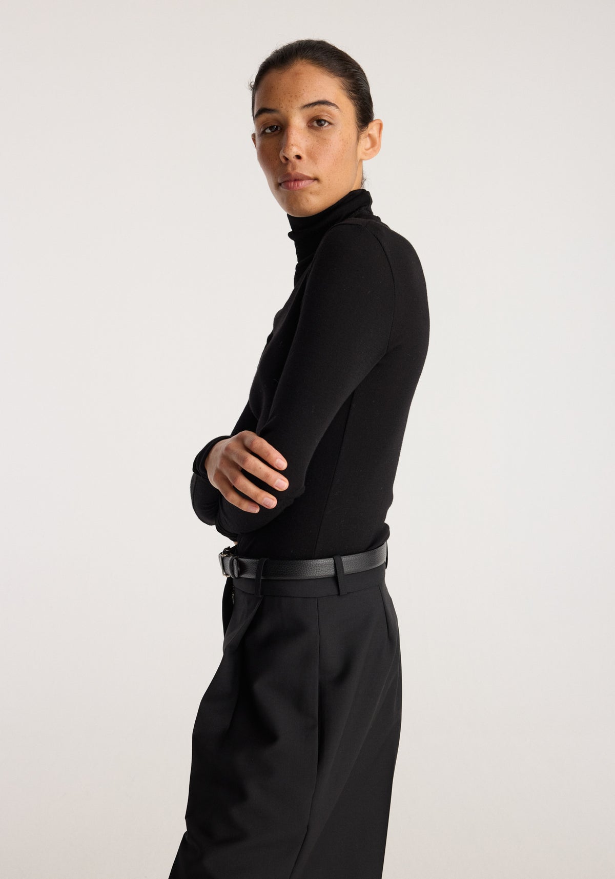 Fine merino turtleneck | black worn by a model in profile with crossed arms, paired with black tailored trousers against a neutral background