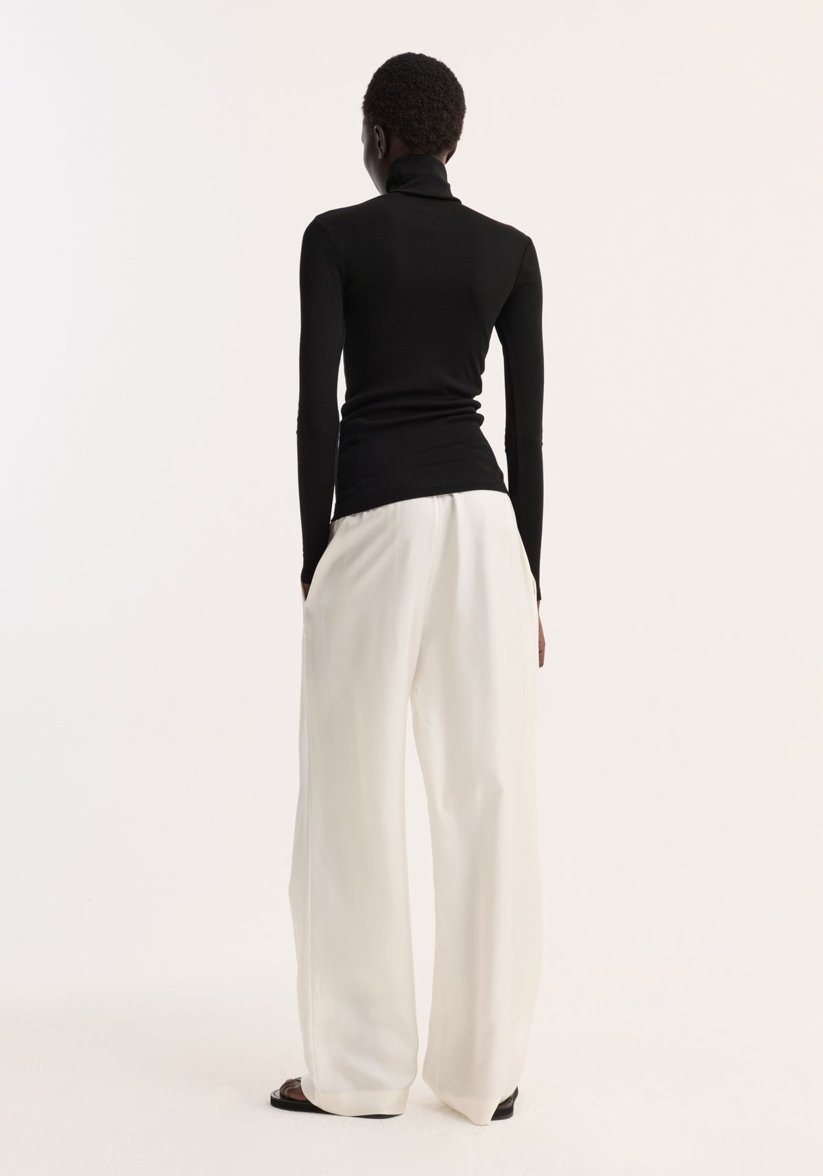 Rear view of a person wearing a fitted black Fine merino turtleneck | black paired with loose white wide-leg pants against a plain light background.