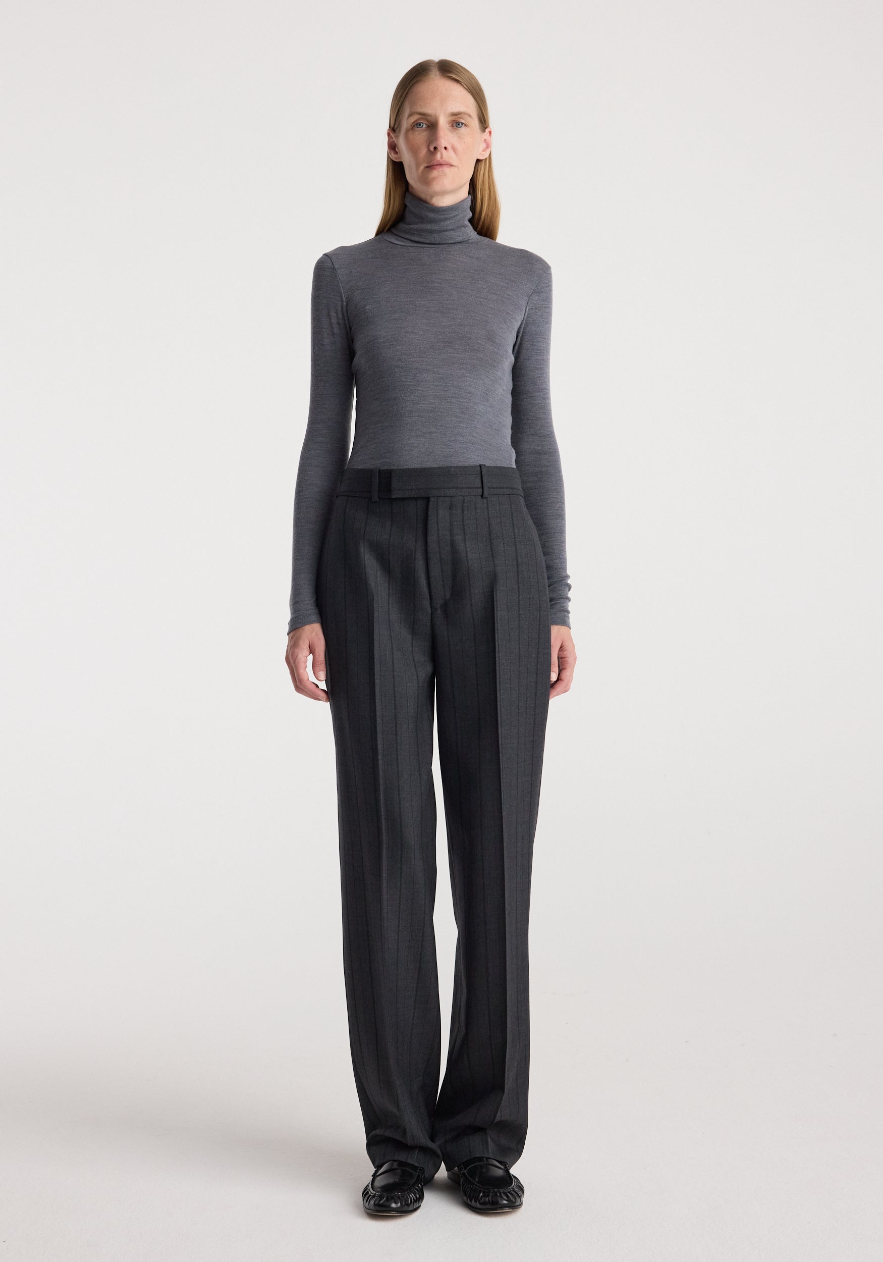 Woman wearing a fitted fine merino turtleneck | mid grey melange paired with dark tailored trousers and black shoes, standing against a plain light background.