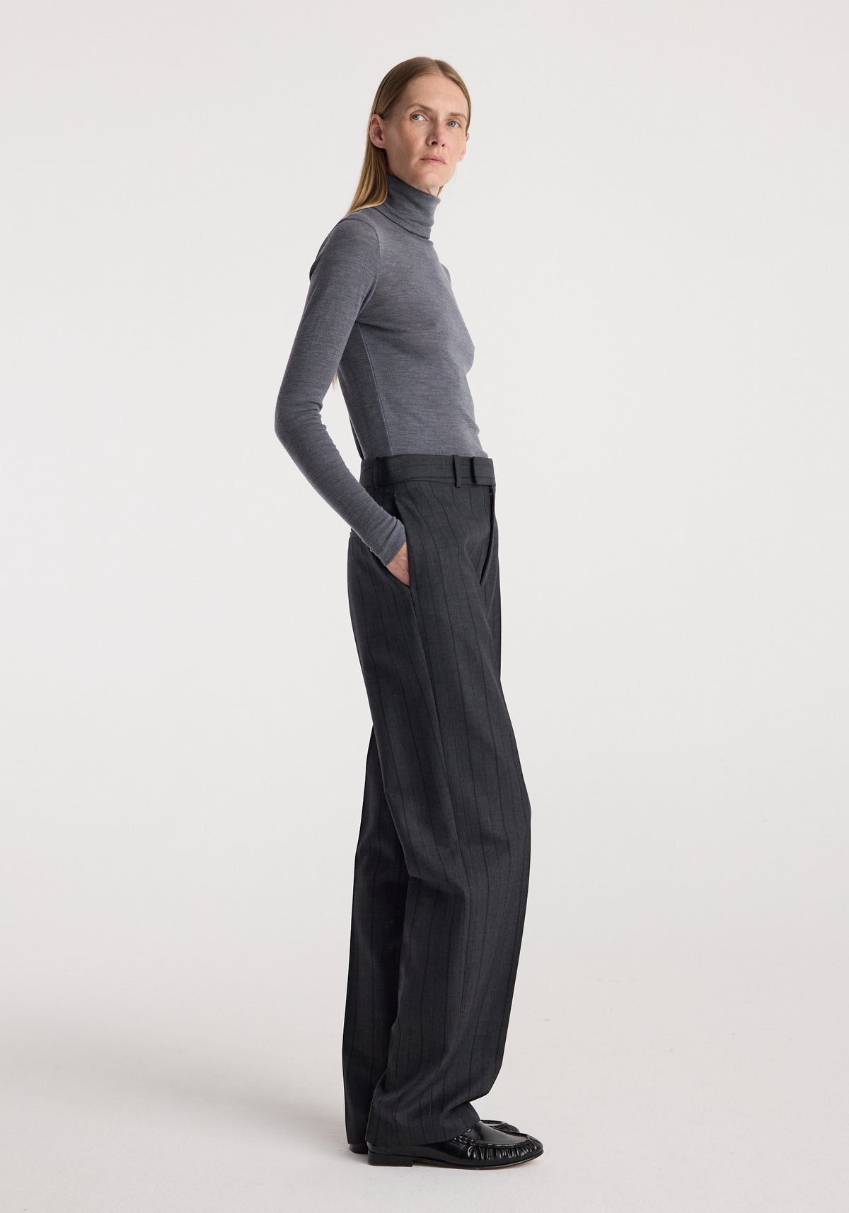 Model wearing a fitted Fine merino turtleneck | mid grey melange with long sleeves and high neckline, styled with loose dark pinstripe trousers in a minimalist studio setting.
