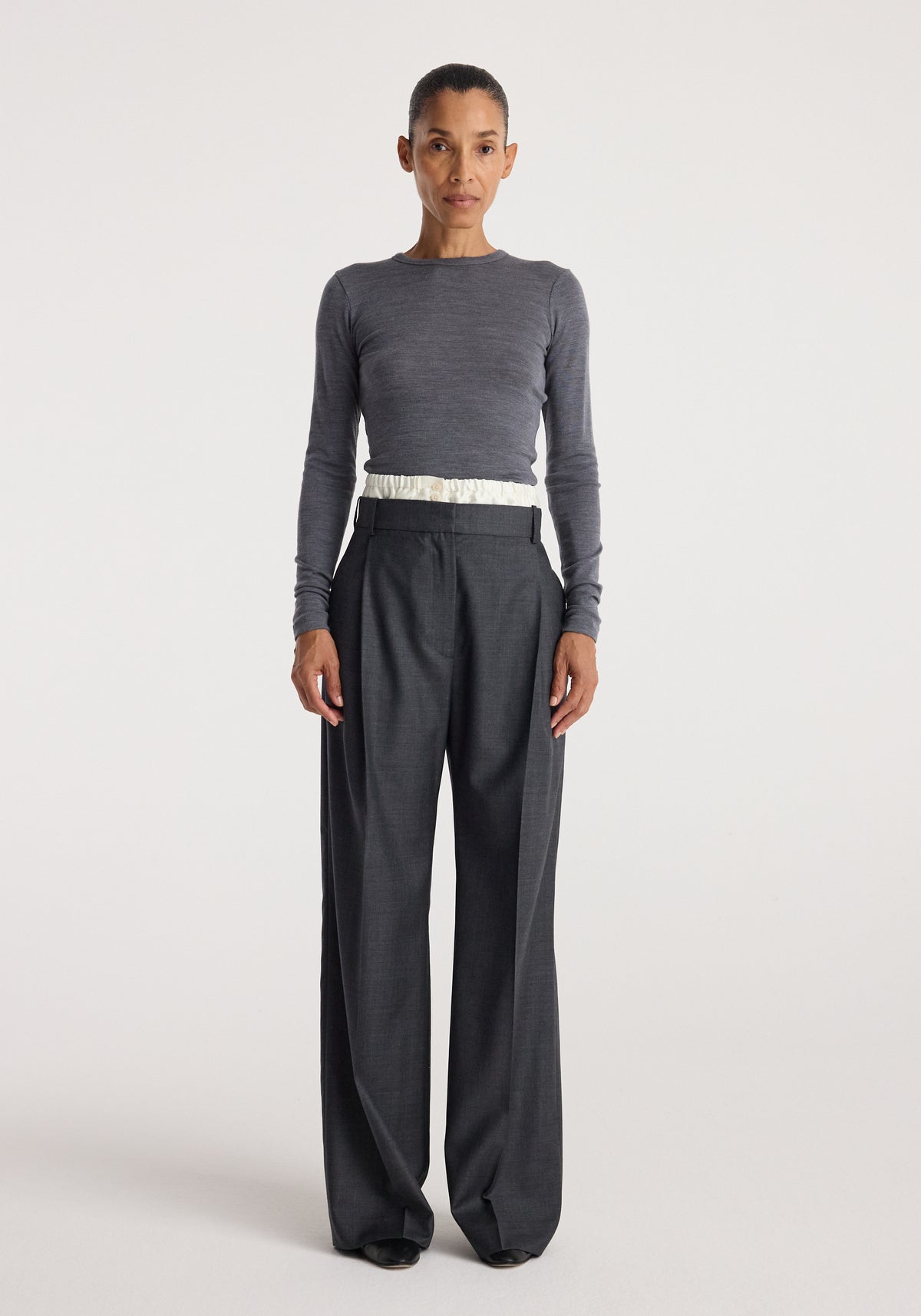 Woman wearing a fitted Fine merino long sleeve | mid grey melange top paired with high-waisted wide-leg dark trousers, standing against a plain light background.