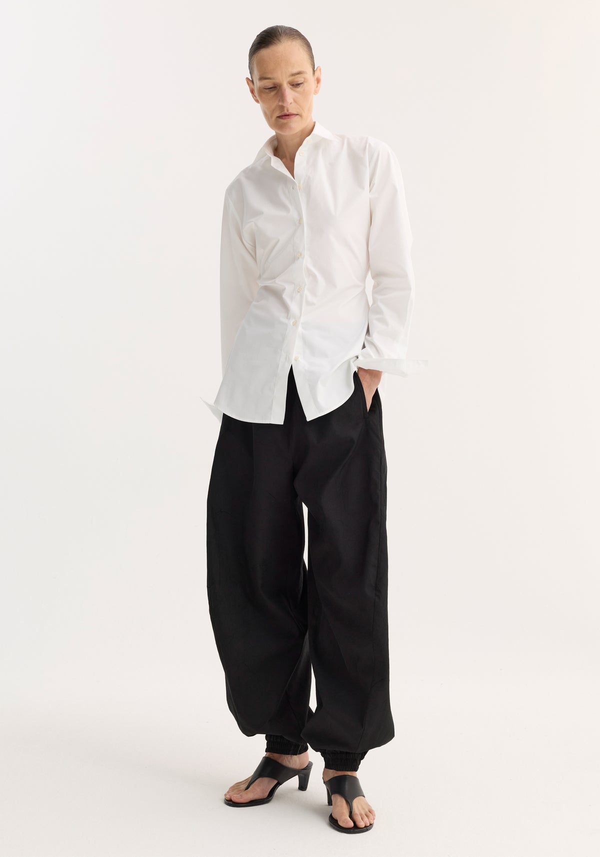 Woman wearing a white Poplin shaped shirt | optic white with long sleeves, paired with loose black pants and black heeled sandals, standing against a plain white background.