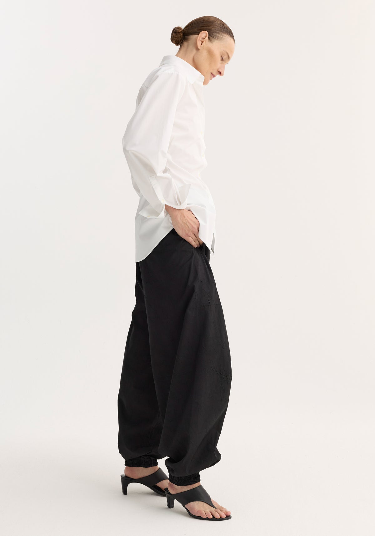 Woman wearing a Poplin shaped shirt | optic white with long sleeves and a classic collar, paired with loose black pants and black heeled sandals, standing in profile.