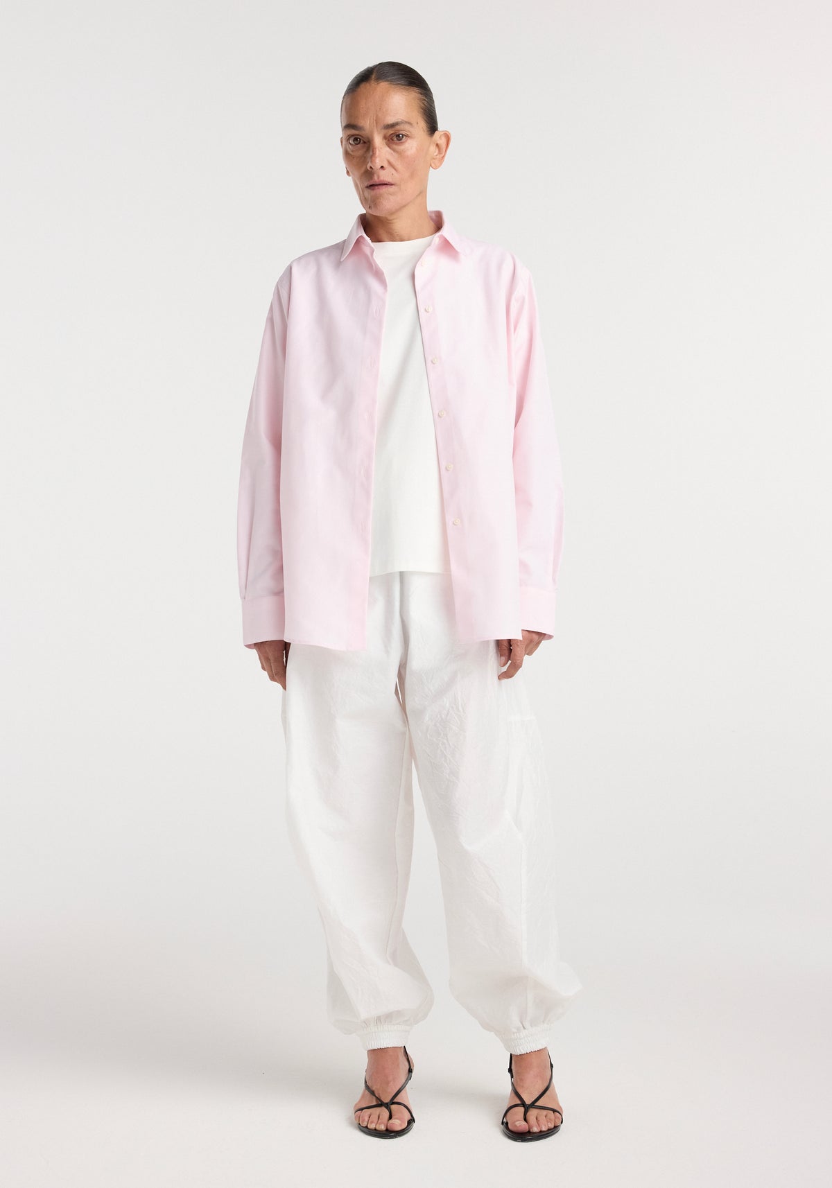 Woman wearing a classic fit shirt | oxford pink, styled open over a white top with loose white pants and black open-toe heels against a plain background.