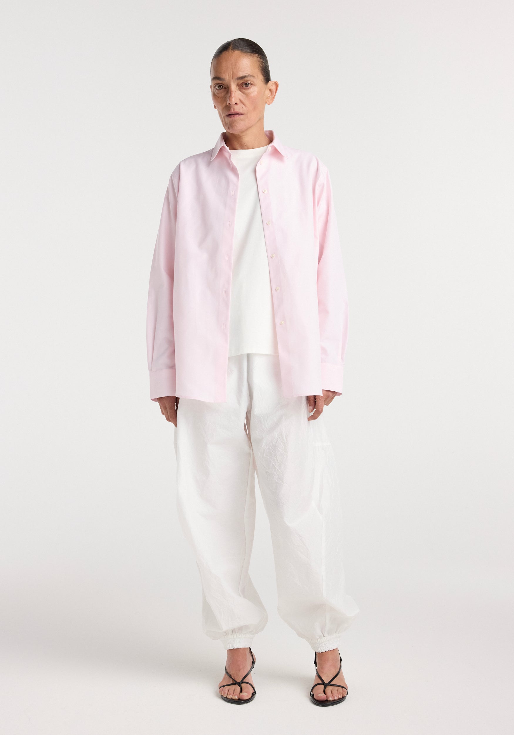 Woman wearing a classic fit shirt | oxford pink, styled open over a white top with loose white pants and black open-toe heels against a plain background.