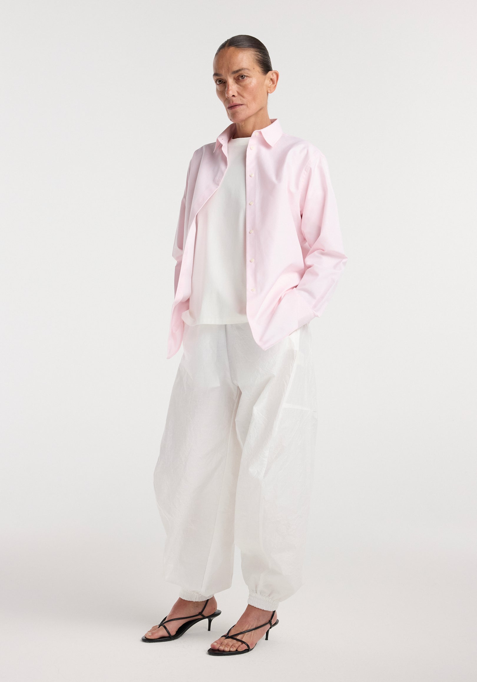 Woman wearing a Classic fit shirt | oxford pink over a white top paired with loose white pants and black heeled sandals, standing against a plain white background.