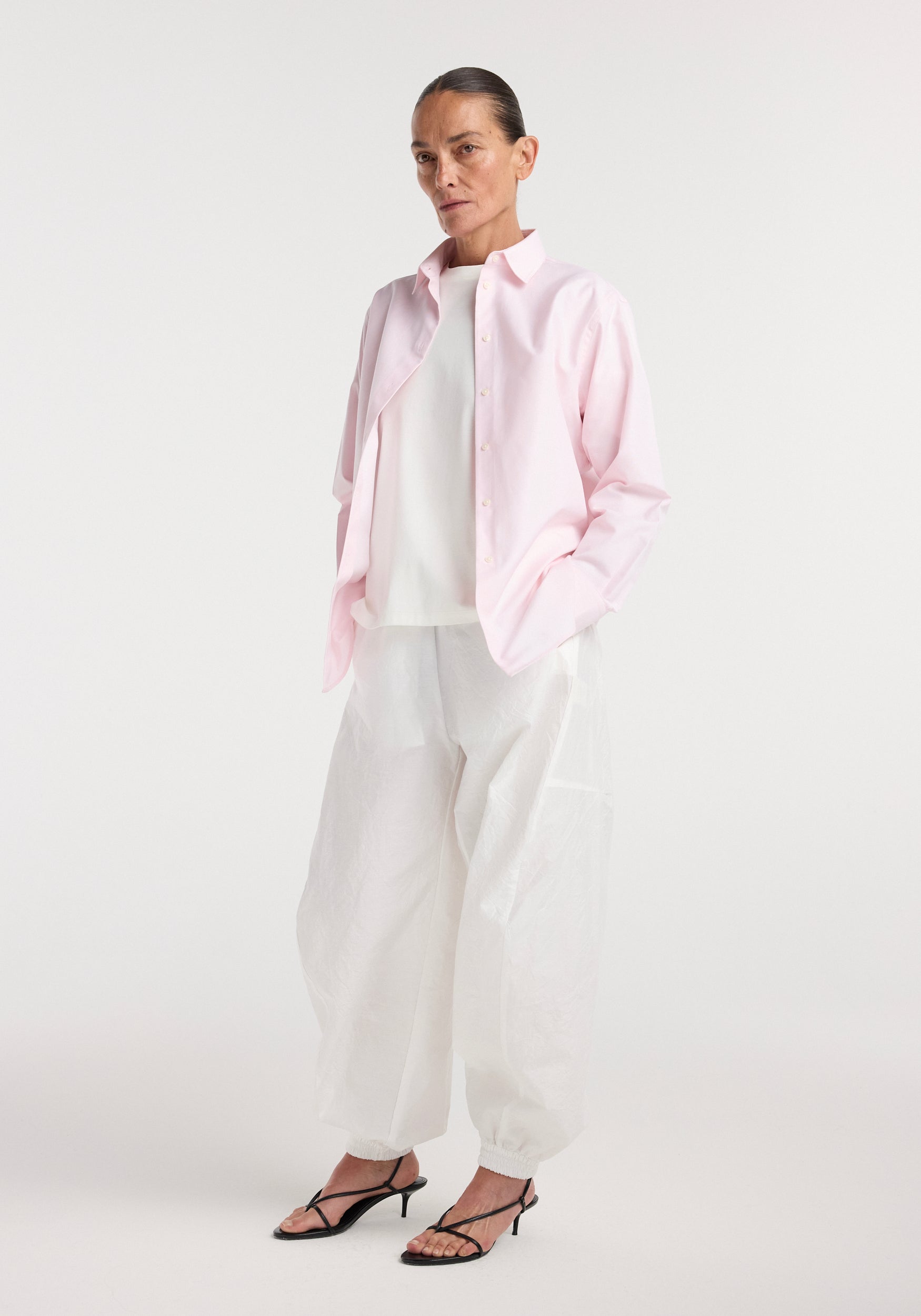 Woman wearing a Classic fit shirt | oxford pink over a white top paired with loose white pants and black heeled sandals, standing against a plain white background.