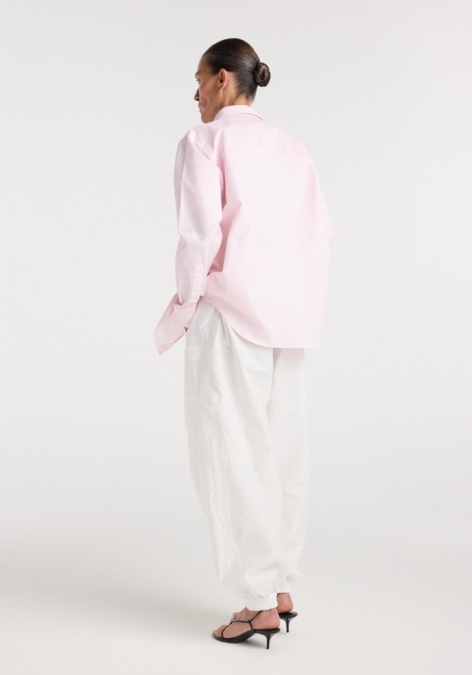 Woman wearing a Classic fit shirt | oxford pink paired with loose white pants and black heeled sandals, standing against a plain white background, viewed from behind.