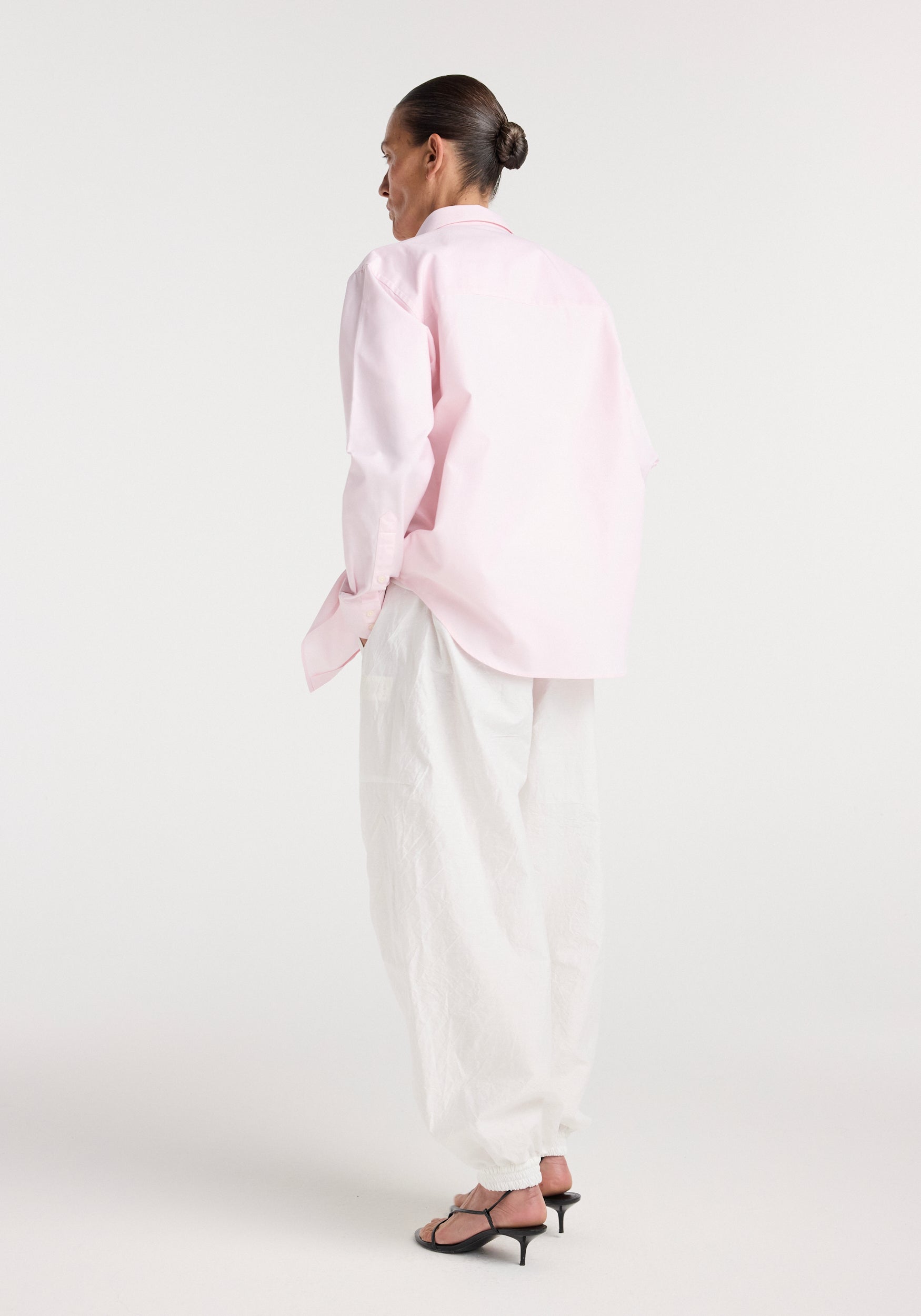Woman wearing a Classic fit shirt | oxford pink paired with loose white pants and black heeled sandals, standing against a plain white background, viewed from behind.