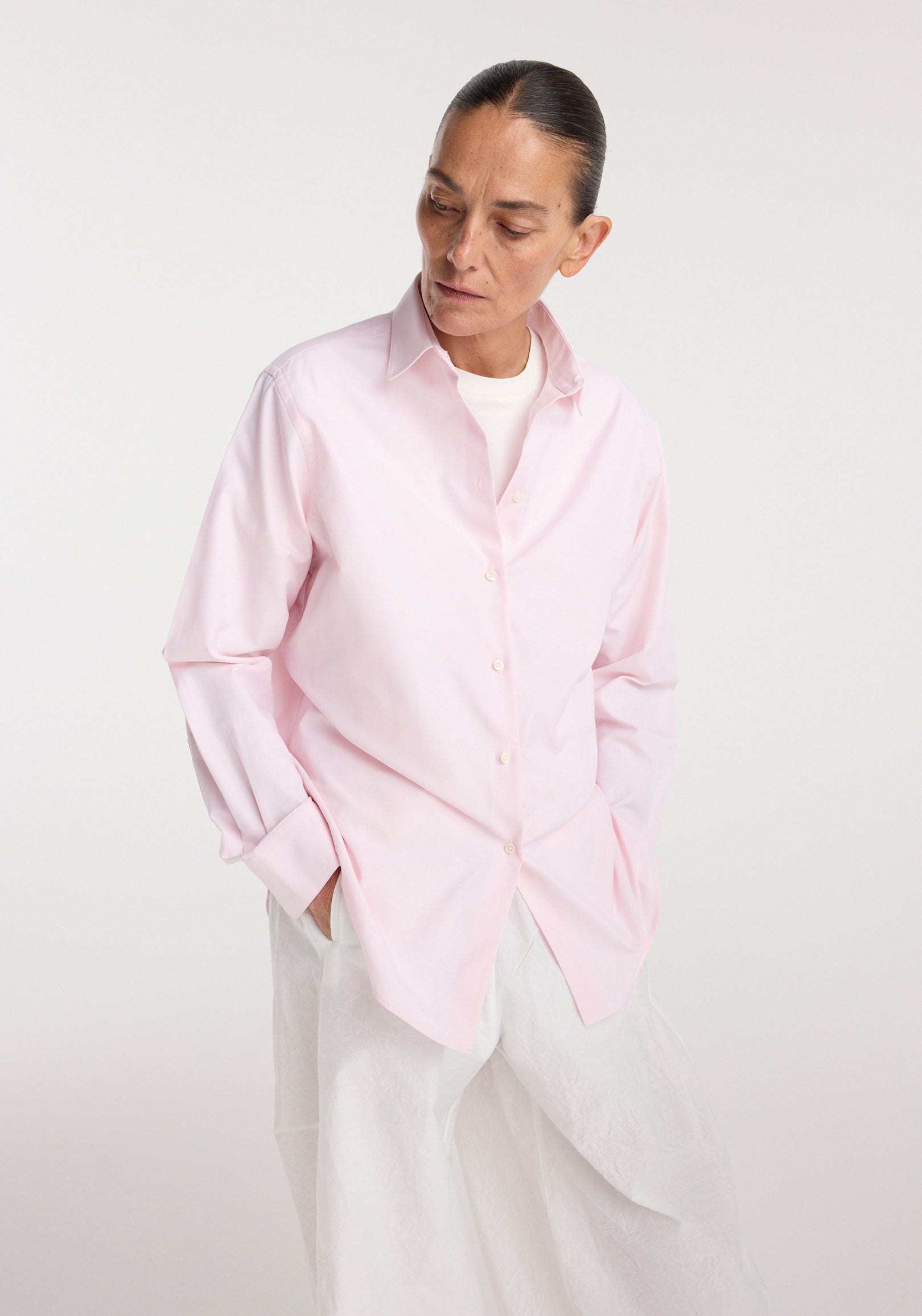 Woman wearing a light pink button-up Classic fit shirt | oxford pink over a white top paired with loose white pants, standing against a plain background with hands in pockets.