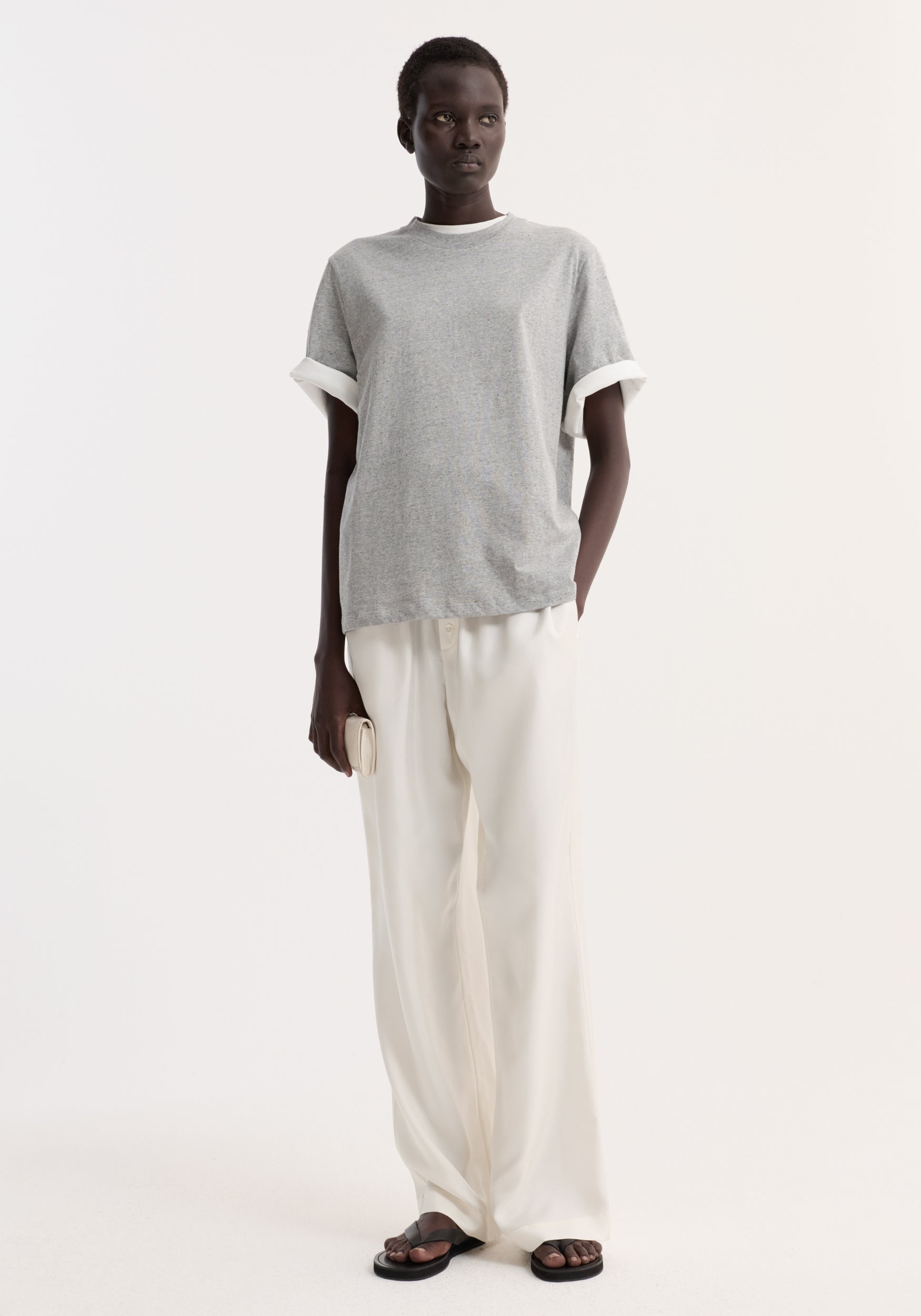 A person wearing a slub melange cotton t-shirt | light grey melange paired with wide-leg white pants and black sandals, standing against a plain light background.