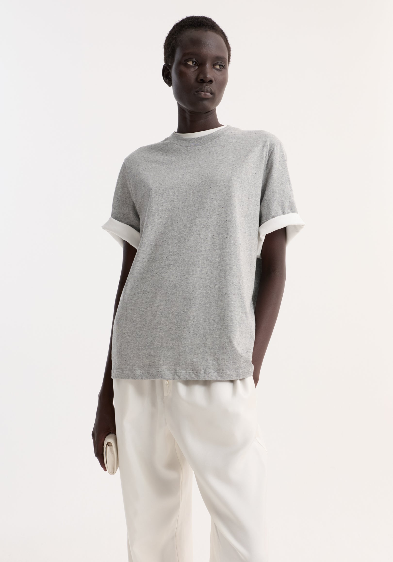 Woman wearing a light grey melange slub melange cotton t-shirt with rolled sleeves, paired with white pants, standing against a plain white background.