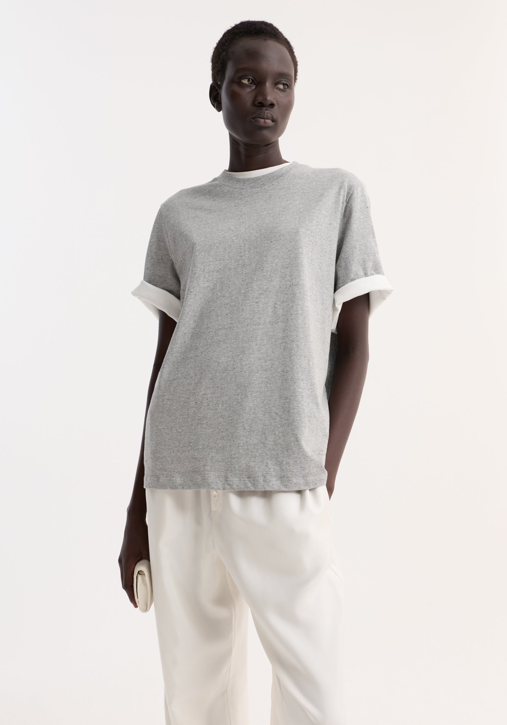 Woman wearing a light grey melange slub melange cotton t-shirt with rolled sleeves, paired with white pants, standing against a plain white background.