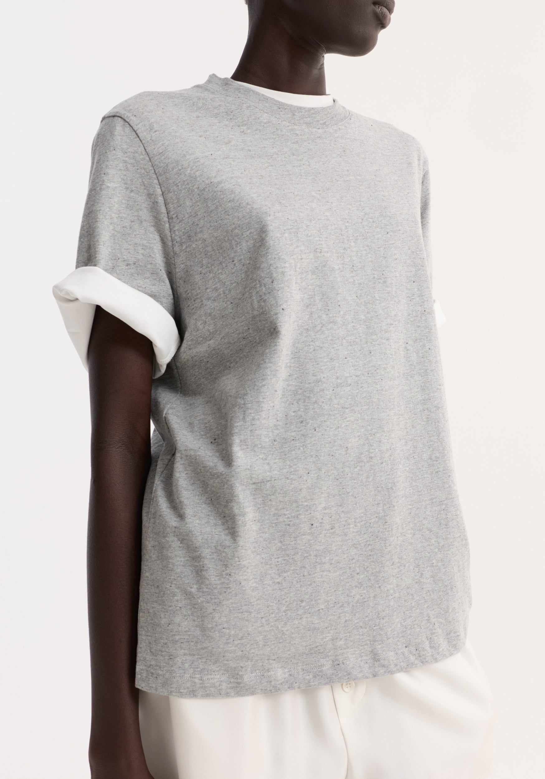 A close-up of a person wearing a Slub melange cotton t-shirt | light grey melange with rolled sleeves, paired with white pants against a plain white background.