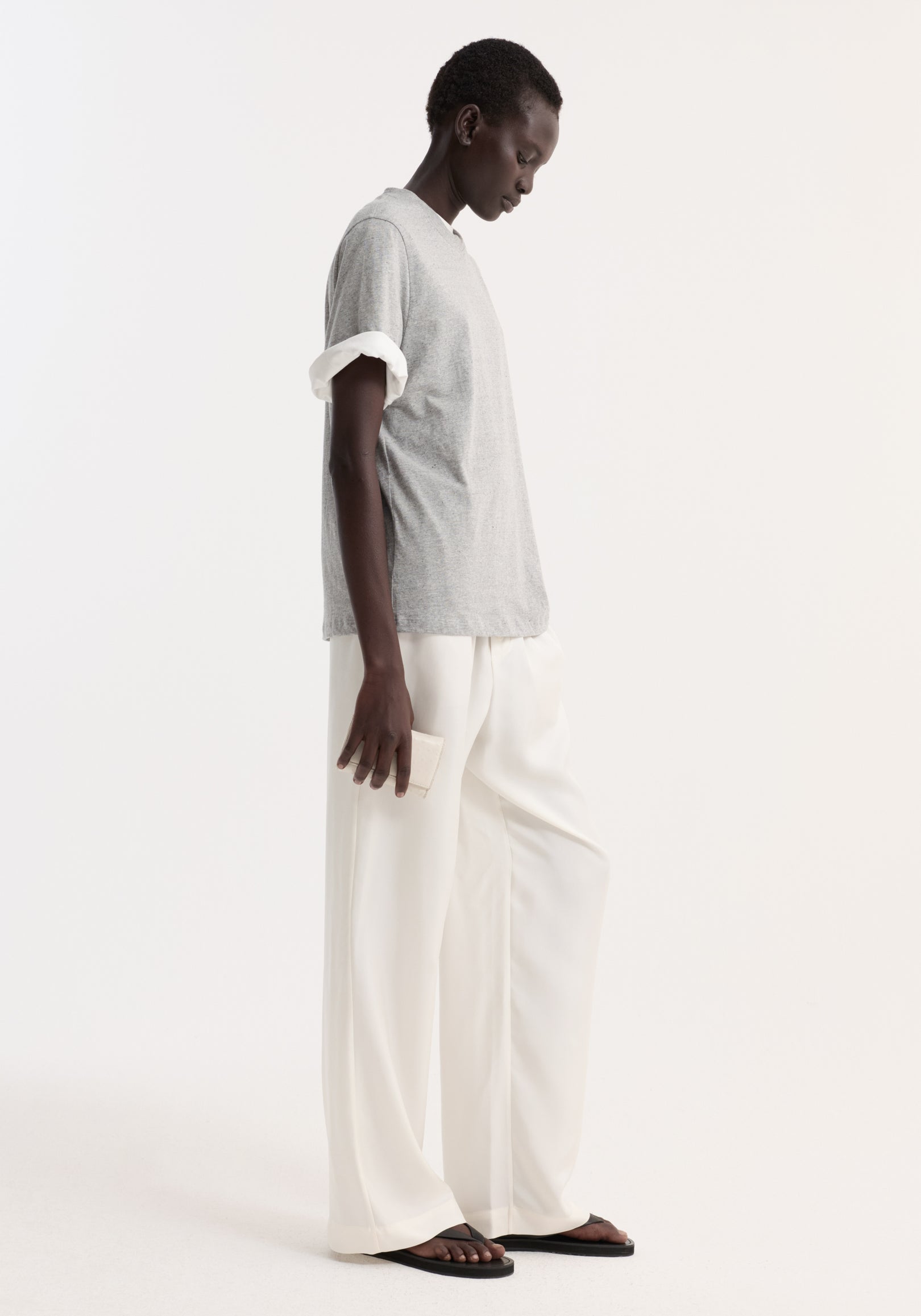 Model wearing a light grey melange slub melange cotton t-shirt with rolled-up sleeves, paired with loose white pants and black sandals, standing against a plain background.
