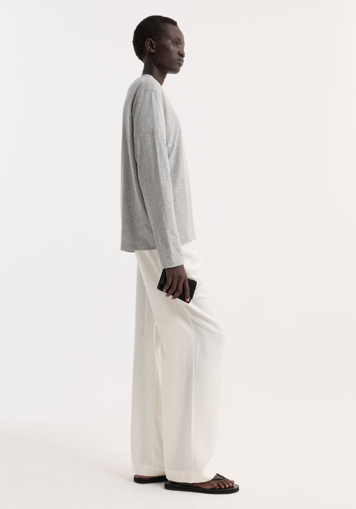 Person in profile wearing a Slub melange cotton long sleeve in light grey melange, paired with loose white trousers and black flat slide sandals, holding a black wallet.