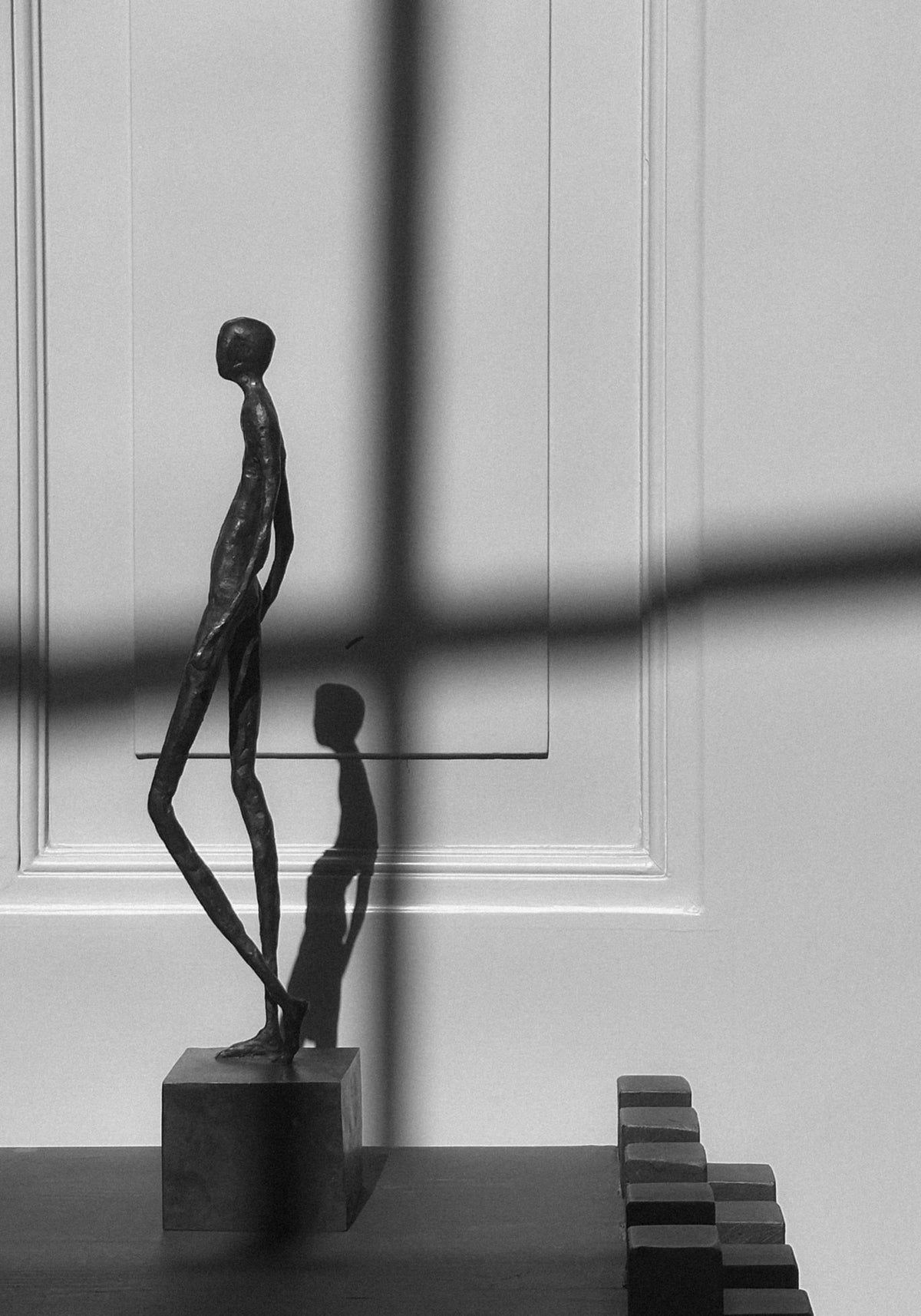 Bronze Sculpture | Quiet Confidence: A slender, handcrafted bronze figure by Veri Wisman on a cubic plinth, highlighted by a cross-shaped shadow on a white paneled background.