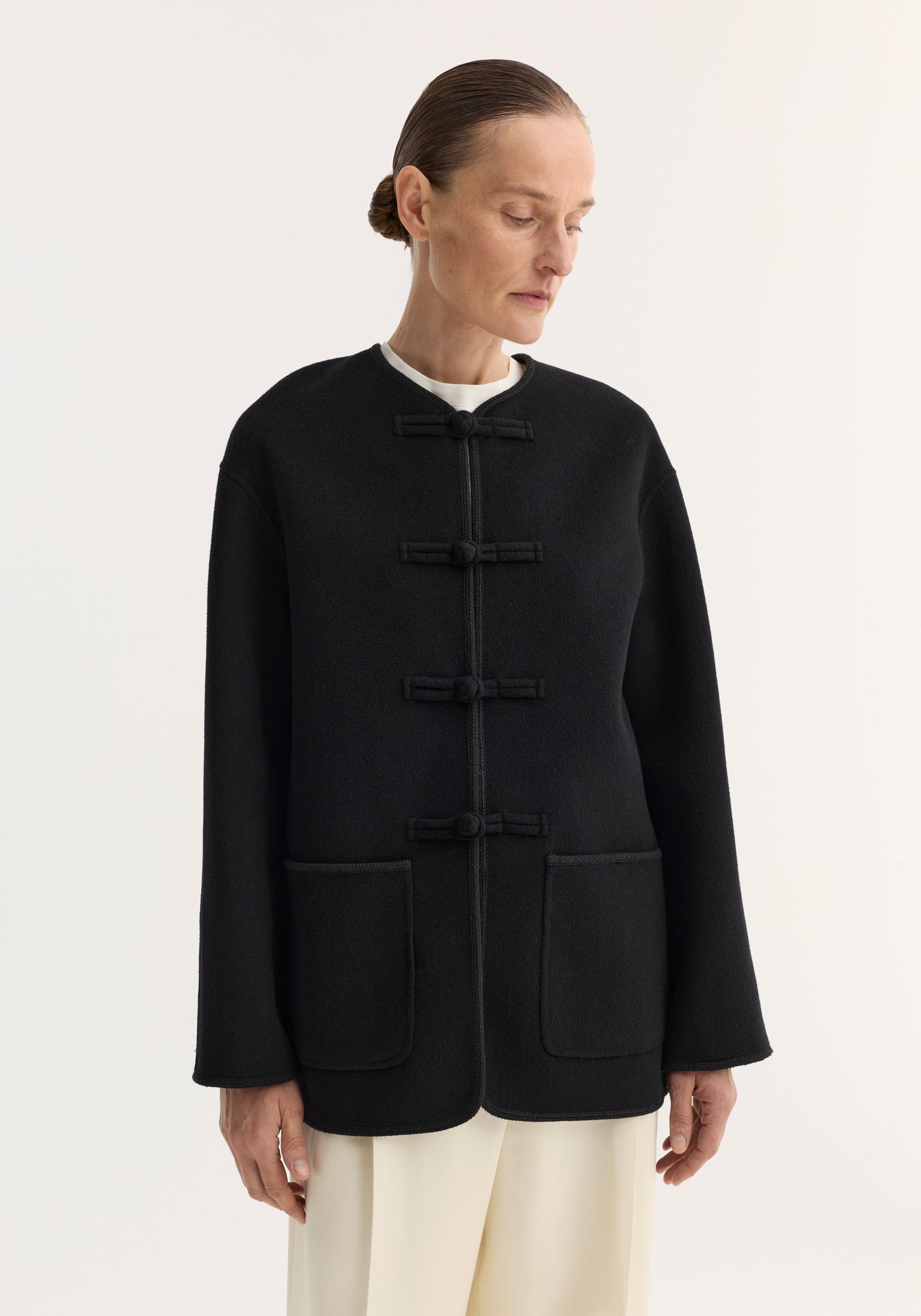 Model in black double-faced wool pankou jacket with frog-style toggles, embroidered edges, and large patch pockets, paired with cream trousers.