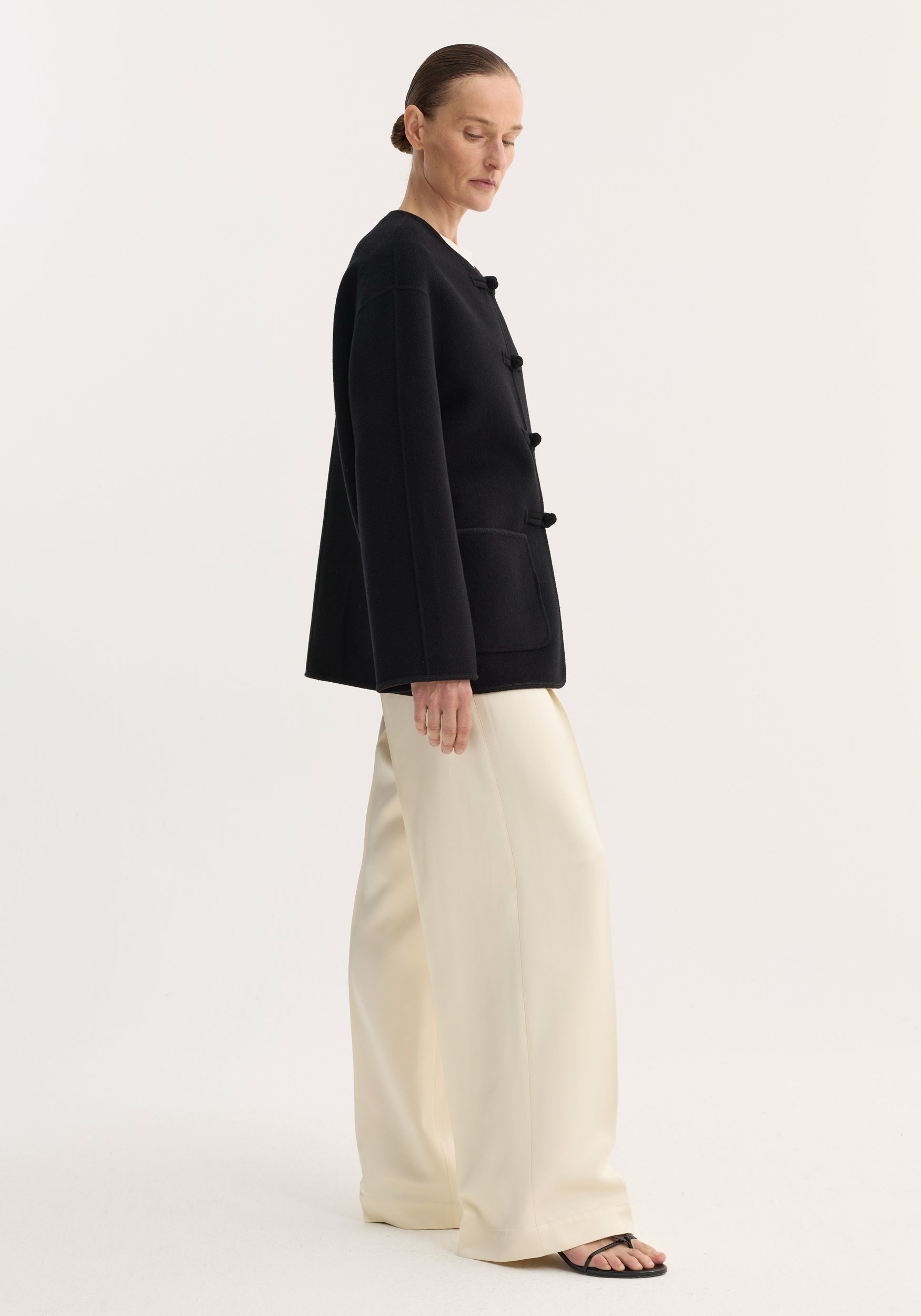 Woman models Double-faced wool pankou jacket with frog-style button closures, embroidered edges, and patch pockets, paired with wide-leg trousers and sandals.