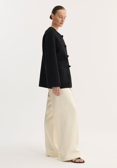 Model wearing the Mandarin double-faced wool jacket in black, featuring frog-style closures, embroidered edges, and patch pockets, paired with wide-leg trousers and slide sandals.
