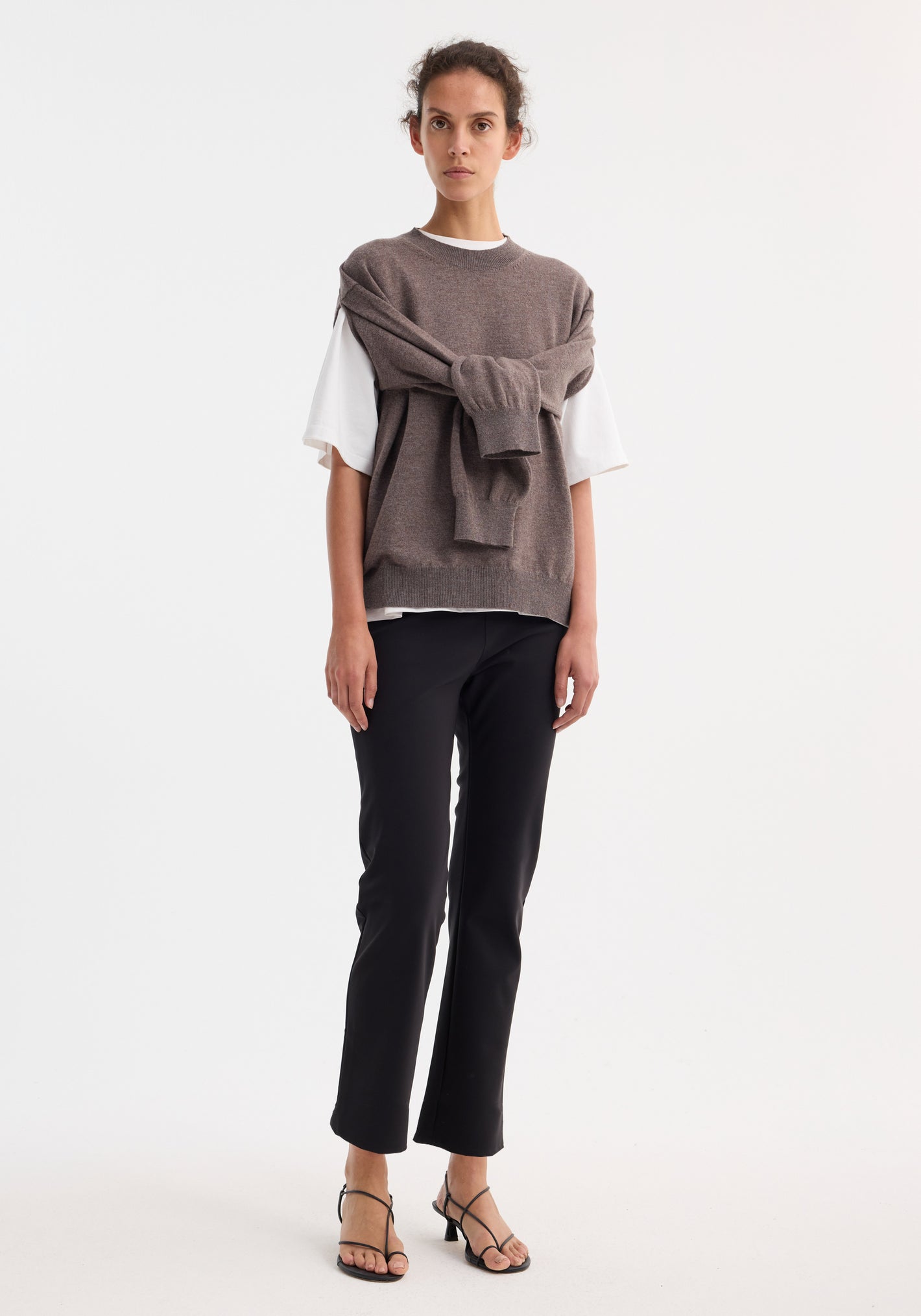Woman wearing cigarette scuba trousers | black, showcasing a tailored ankle-length silhouette with a sleek cigarette leg design, paired with a taupe-gray knit sleeveless sweater.