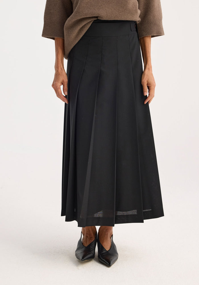 Person wearing a Plissé wrap skirt, black, featuring pleats, wrap design, and belt detail, paired with a taupe sweater and pointed-toe shoes.