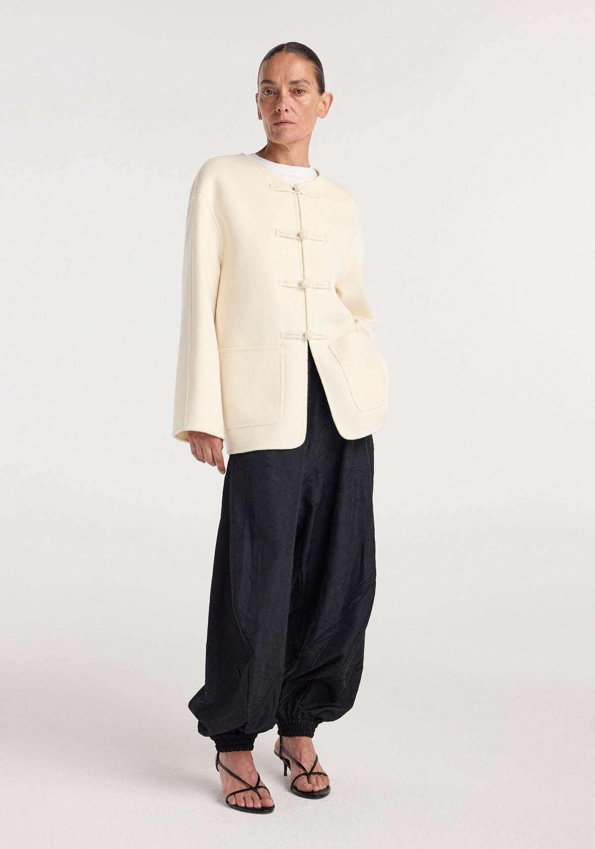 Woman wearing a double-faced wool pankou jacket in off white with traditional button closures, paired with loose black pants and strappy heels, standing against a plain background.