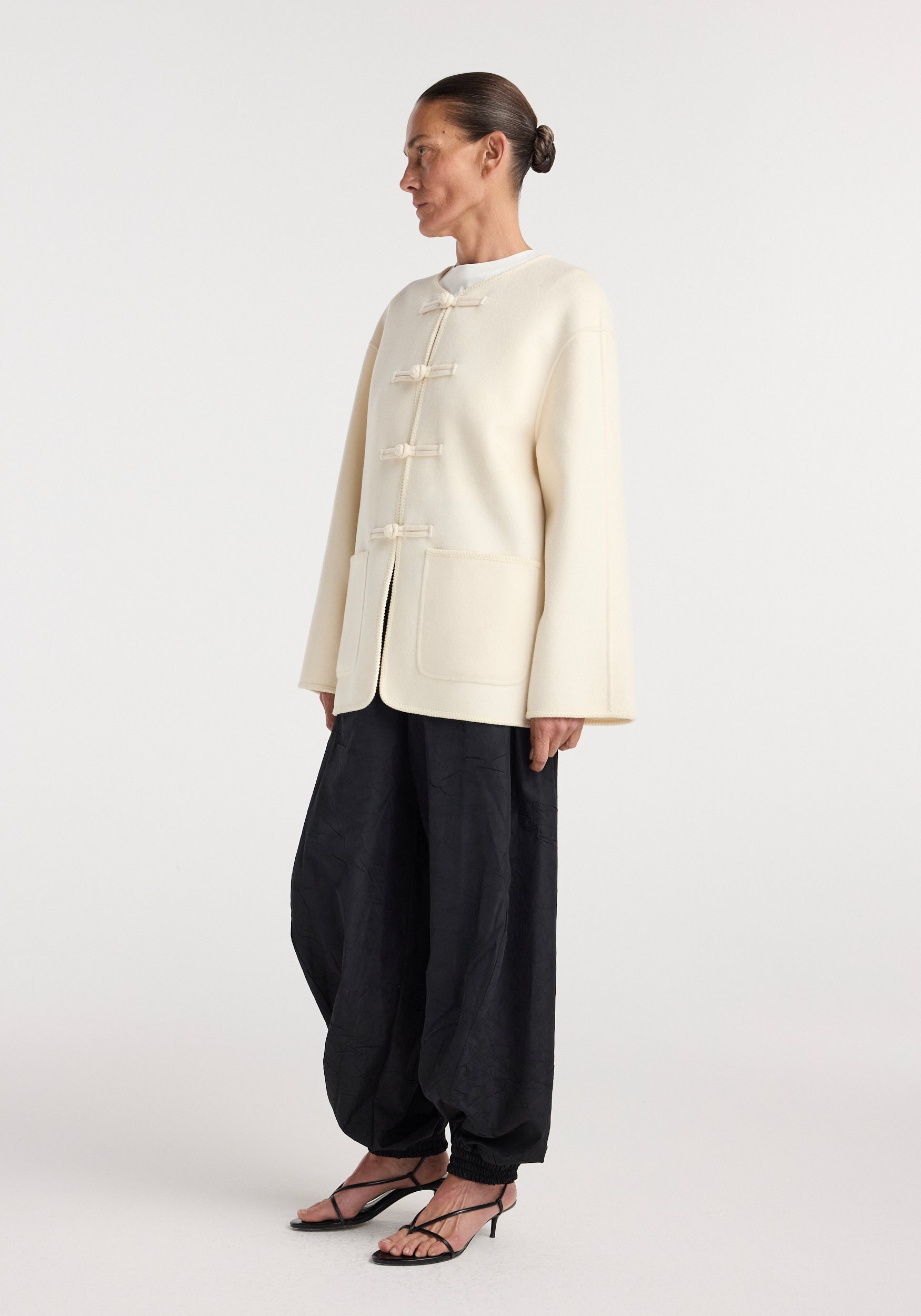 Woman wearing an off-white double-faced wool pankou jacket with traditional knotted closures, paired with loose black pants and strappy heels, standing against a plain background.