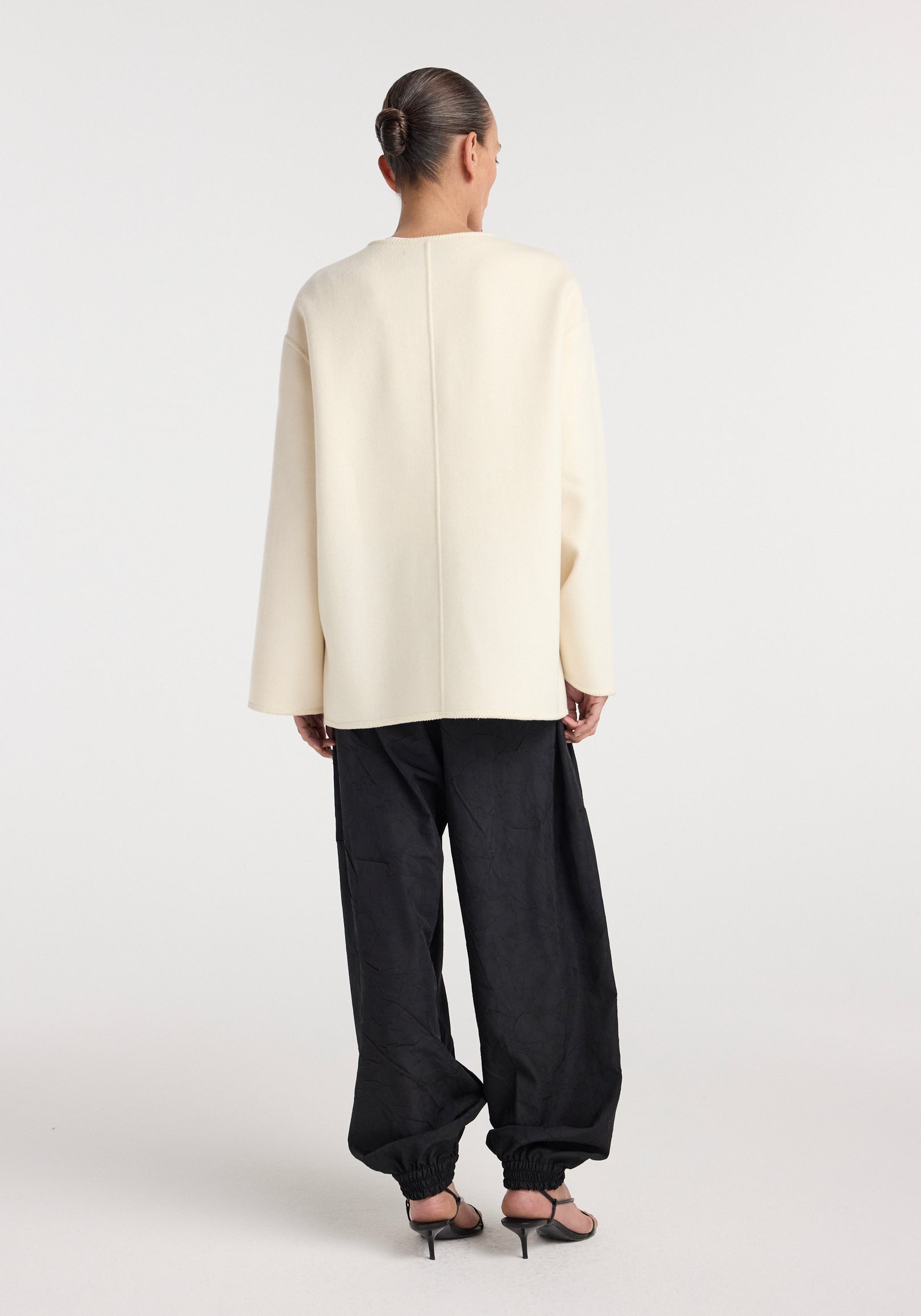 Woman seen from behind wearing an off white Double-faced wool pankou jacket with long sleeves, paired with loose black pants and black heeled sandals against a plain background.