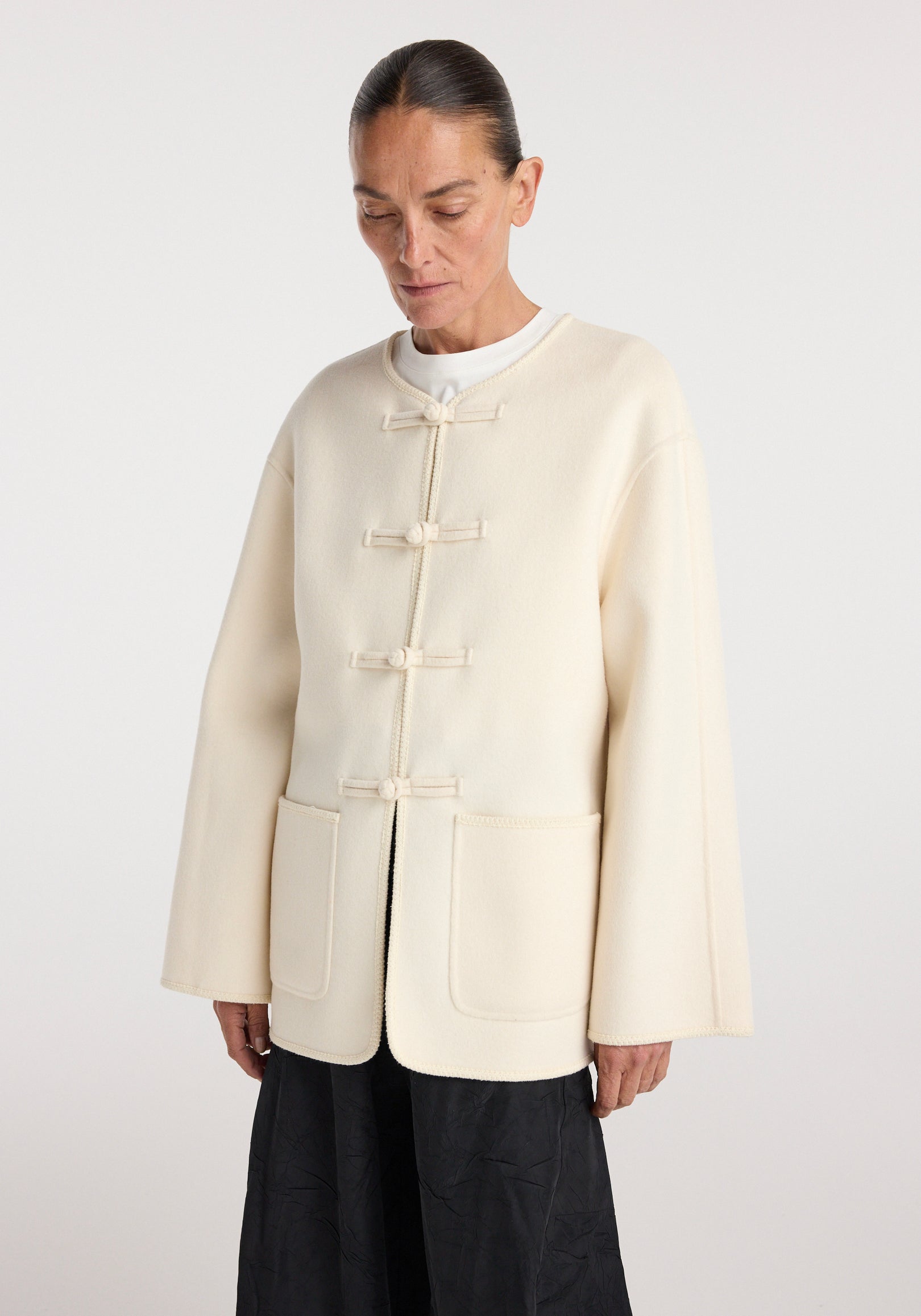 Woman wearing an off white Double-faced wool pankou jacket | off white with traditional pankou knot fastenings and two front pockets over a white top and black skirt.