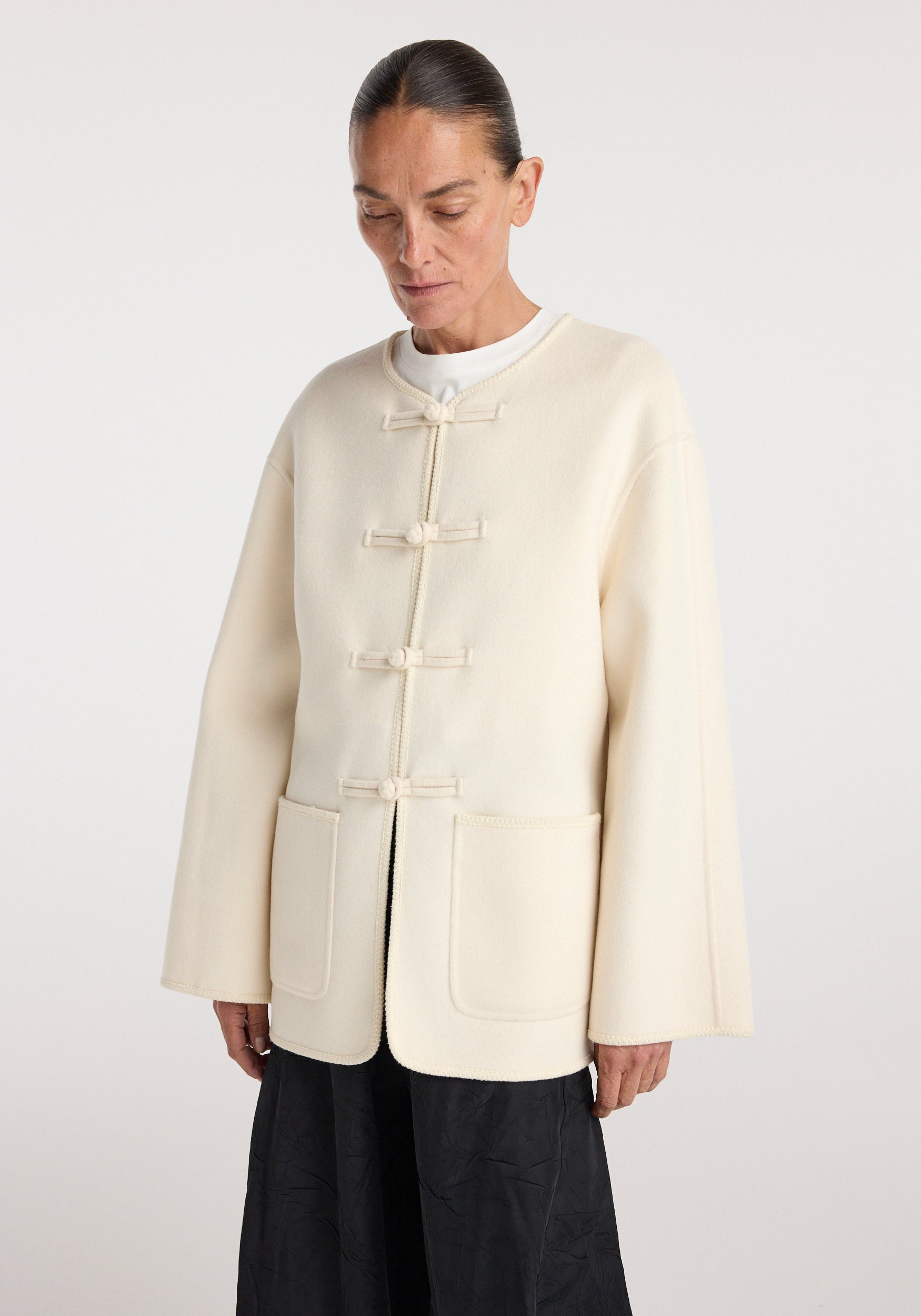 Woman wearing an off white Double-faced wool pankou jacket | off white with traditional pankou knot fastenings and two front pockets over a white top and black skirt.