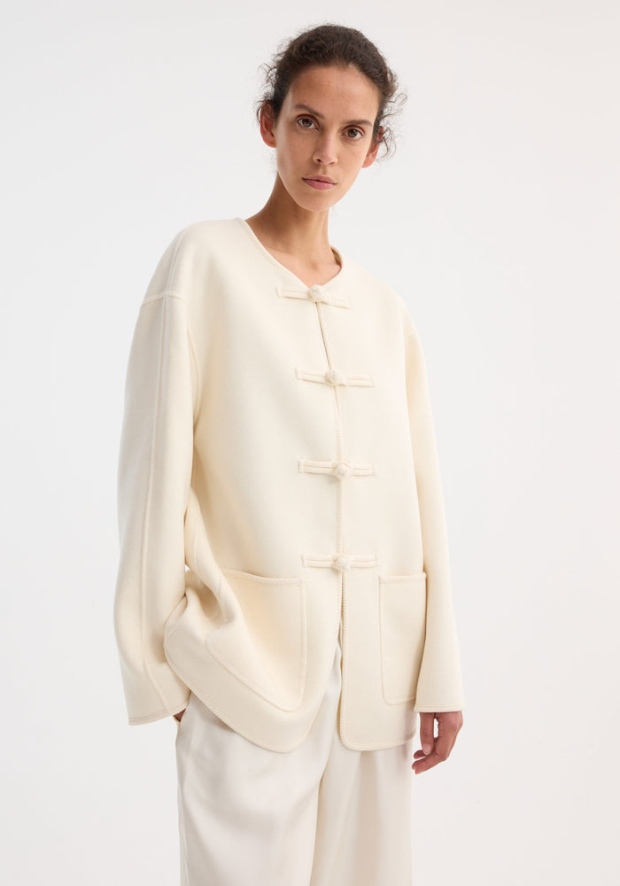 Model wearing Mandarin double-faced wool jacket in off-white, featuring a round neckline, frog-style toggles, two patch pockets, and embroidered edges.